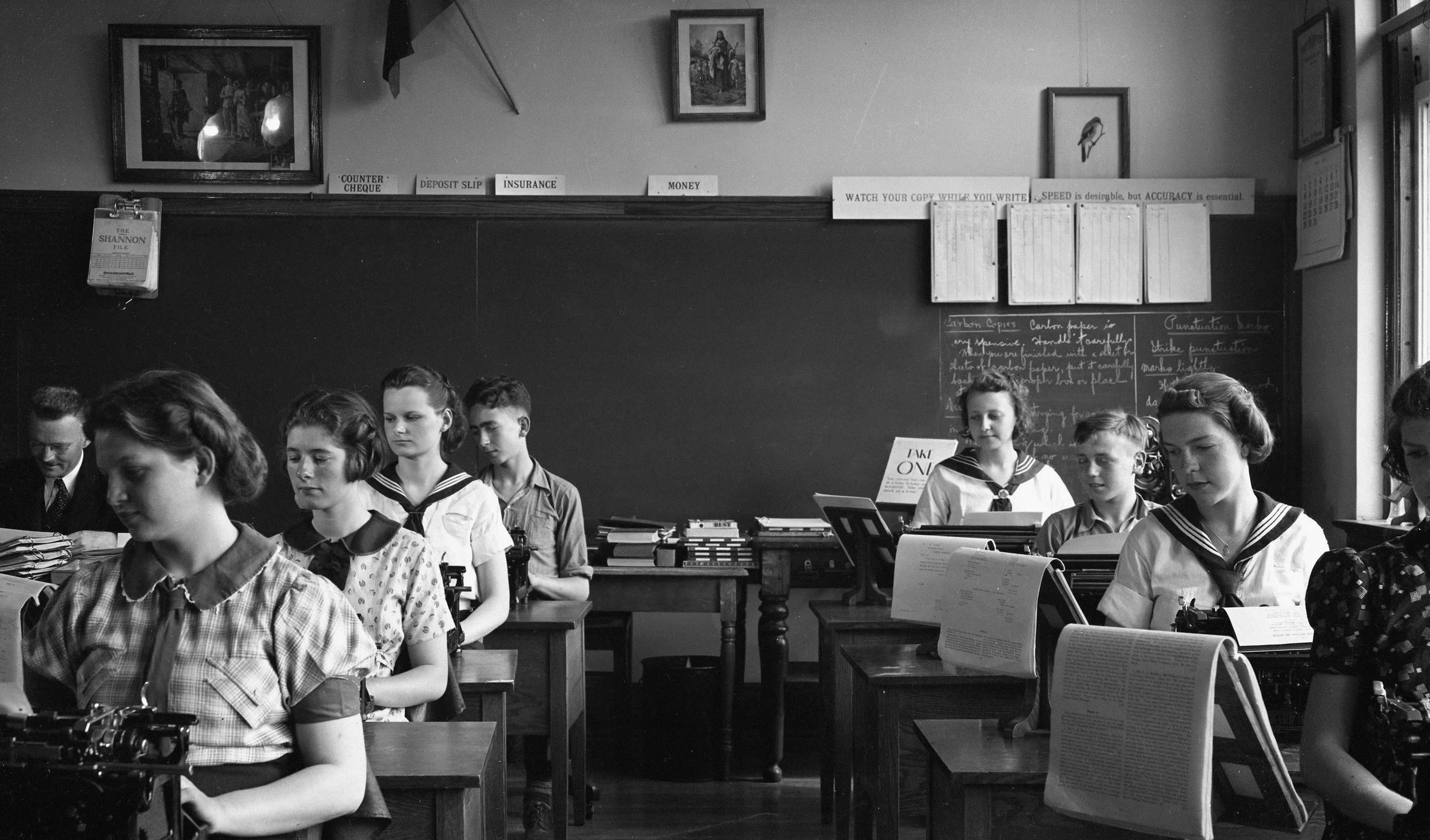 Black-and-white vintage typing class: students at typewriters, a teacher at left, a Good Shepherd picture above the board.
