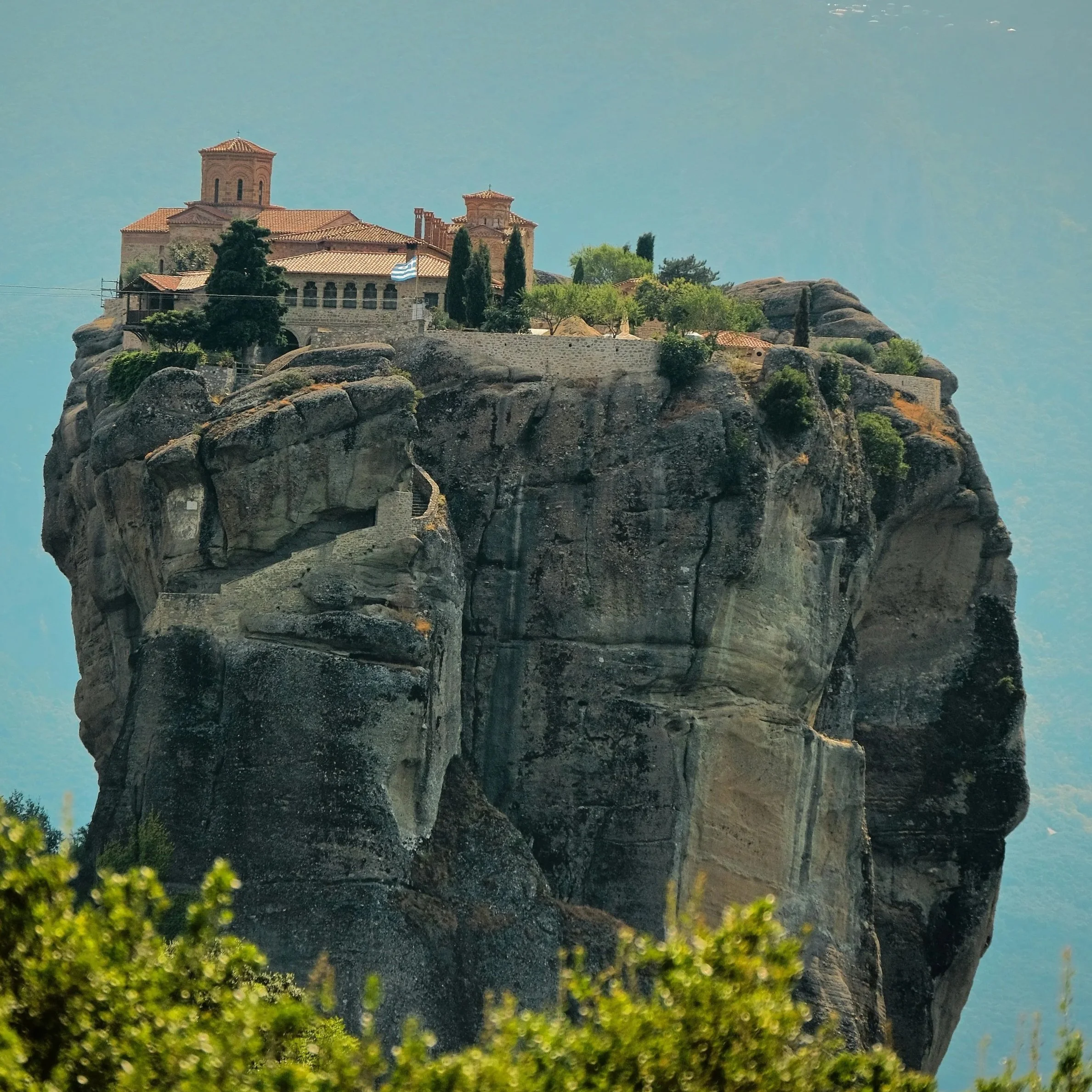 Historic monastery perched on a towering rock formation in Meteora, Greece, with misty mountains and clouds in the background.