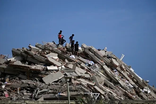 People search through the debris of a collapsed building after an earthquake, with broken concrete slabs and rubble piled beneath a clear blue sky.