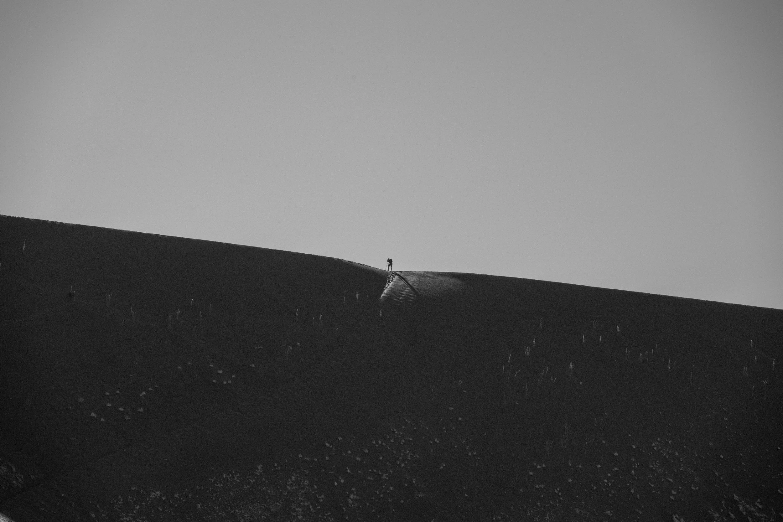 Minimalist black-and-white photograph of a lone person standing atop a dark, sloping ridge against an expansive sky.