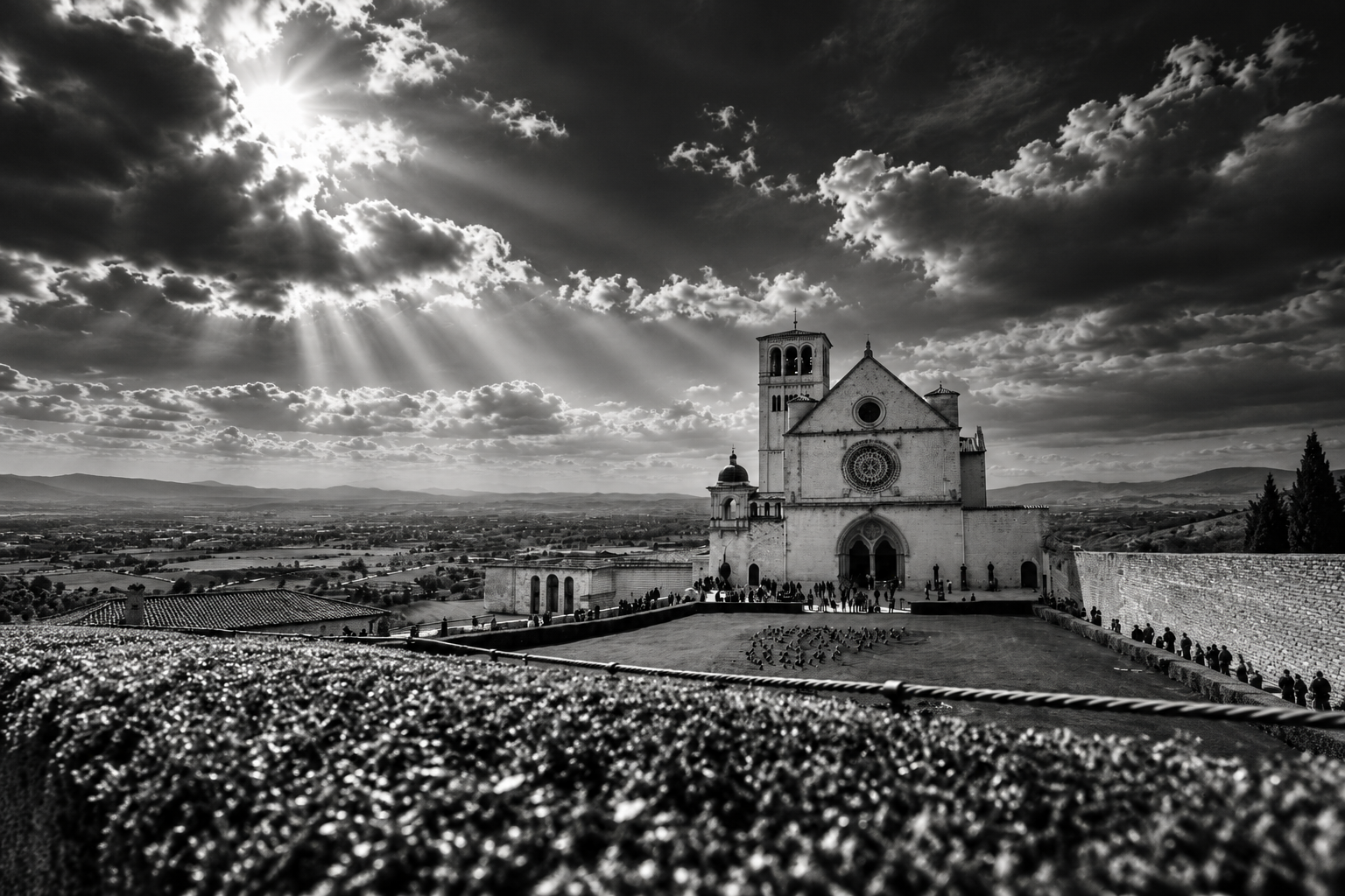Dramatic black-and-white view of the Basilica of Saint Francis in Assisi beneath clouds pierced by sunlight.