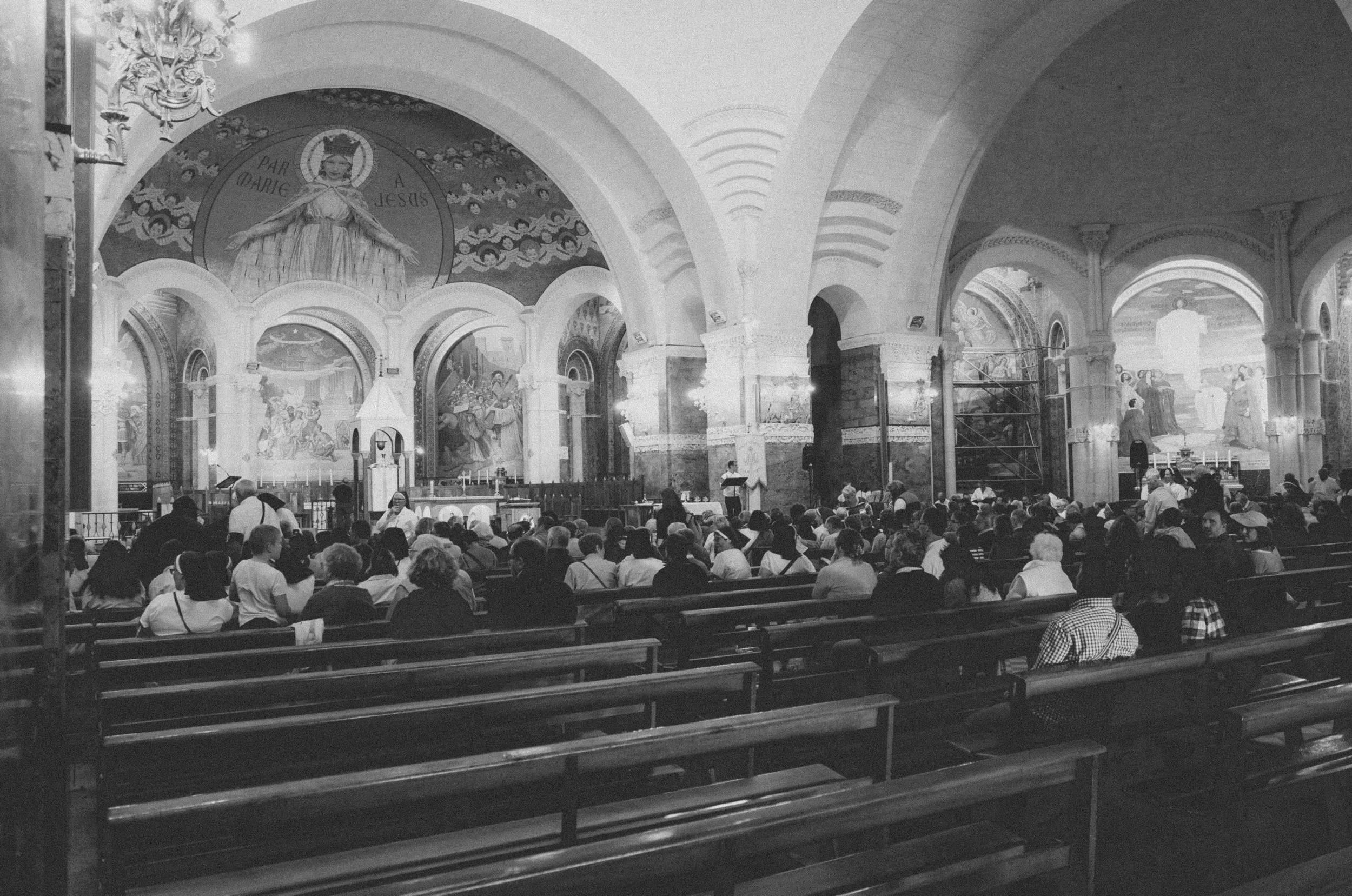 Black-and-white view of worshippers and nuns filling pews in an arched basilica beneath a mosaic of Mary with outstretched arms