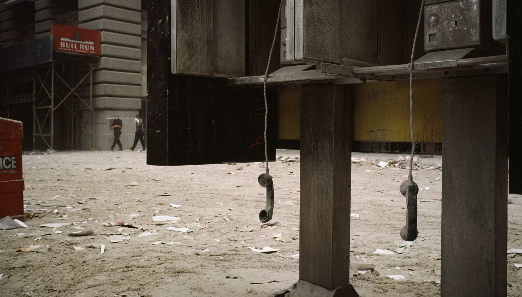 Dust-covered payphone bank with dangling receivers on a debris-filled Lower Manhattan street following the September 11 attacks, with two people walking in the background.