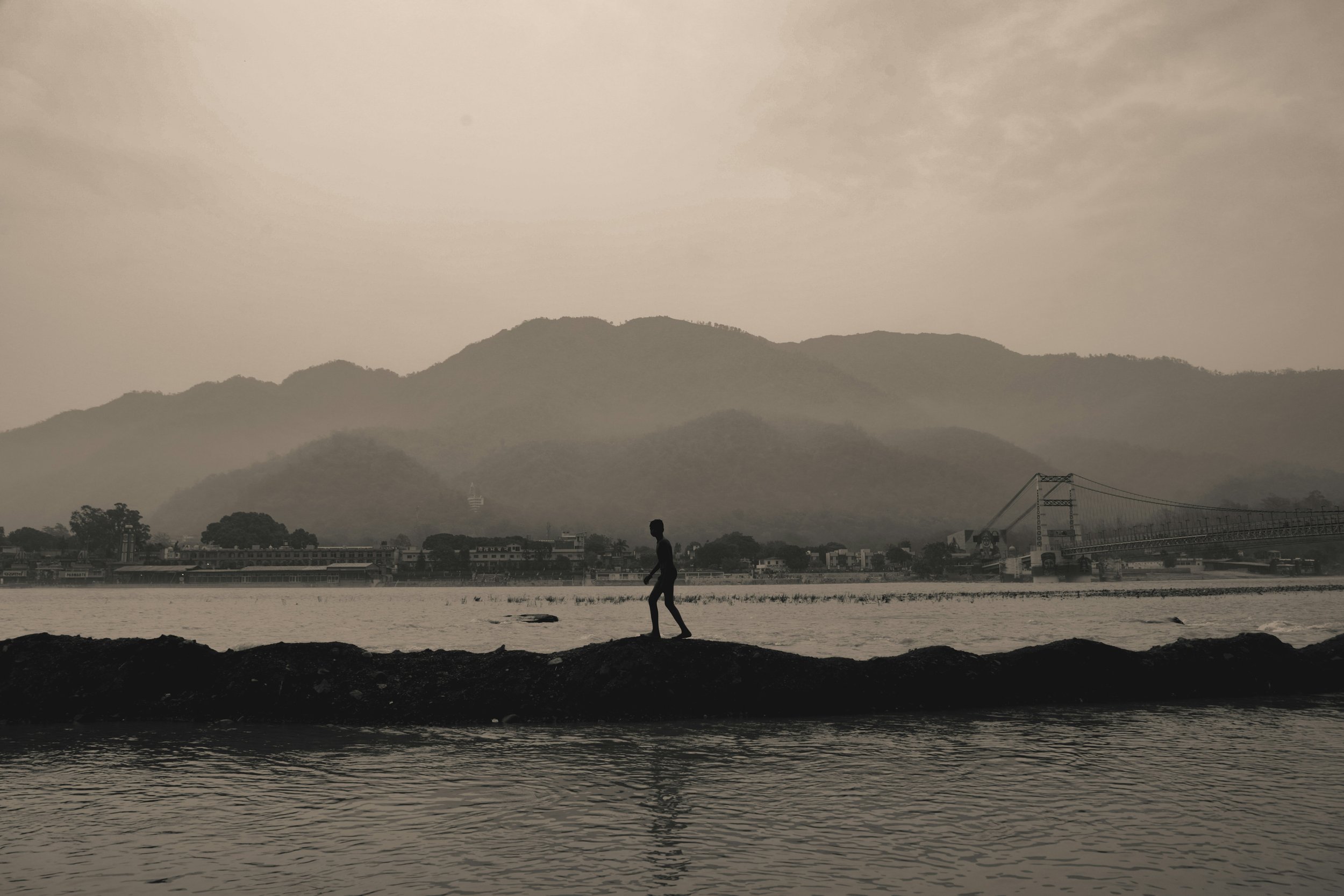 Sepia silhouette of a young man walking barefoot along a riverbank ridge, with misty mountains and a bridge beyond.