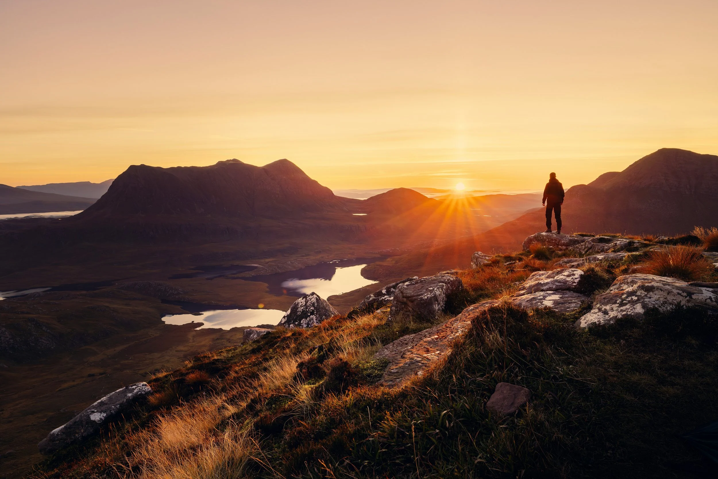 Lone person standing on a rocky mountain ridge at sunrise, looking over glowing valleys and lakes, symbolizing the search for meaning and the seekers’ path.