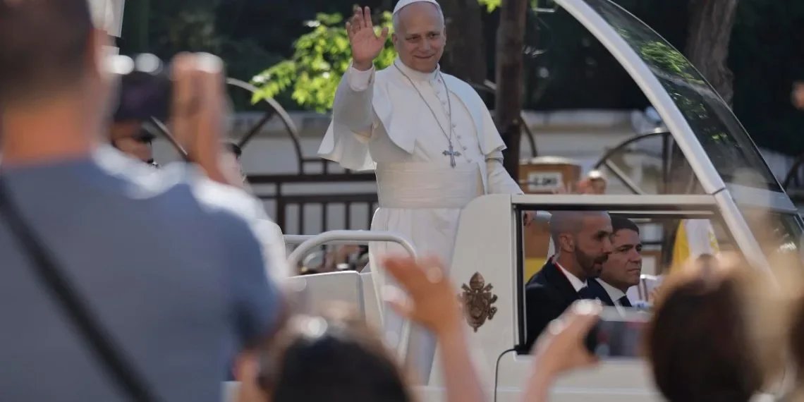 The Pope waves from the popemobile during an outdoor public appearance, surrounded by a crowd of people taking photos and greeting him.