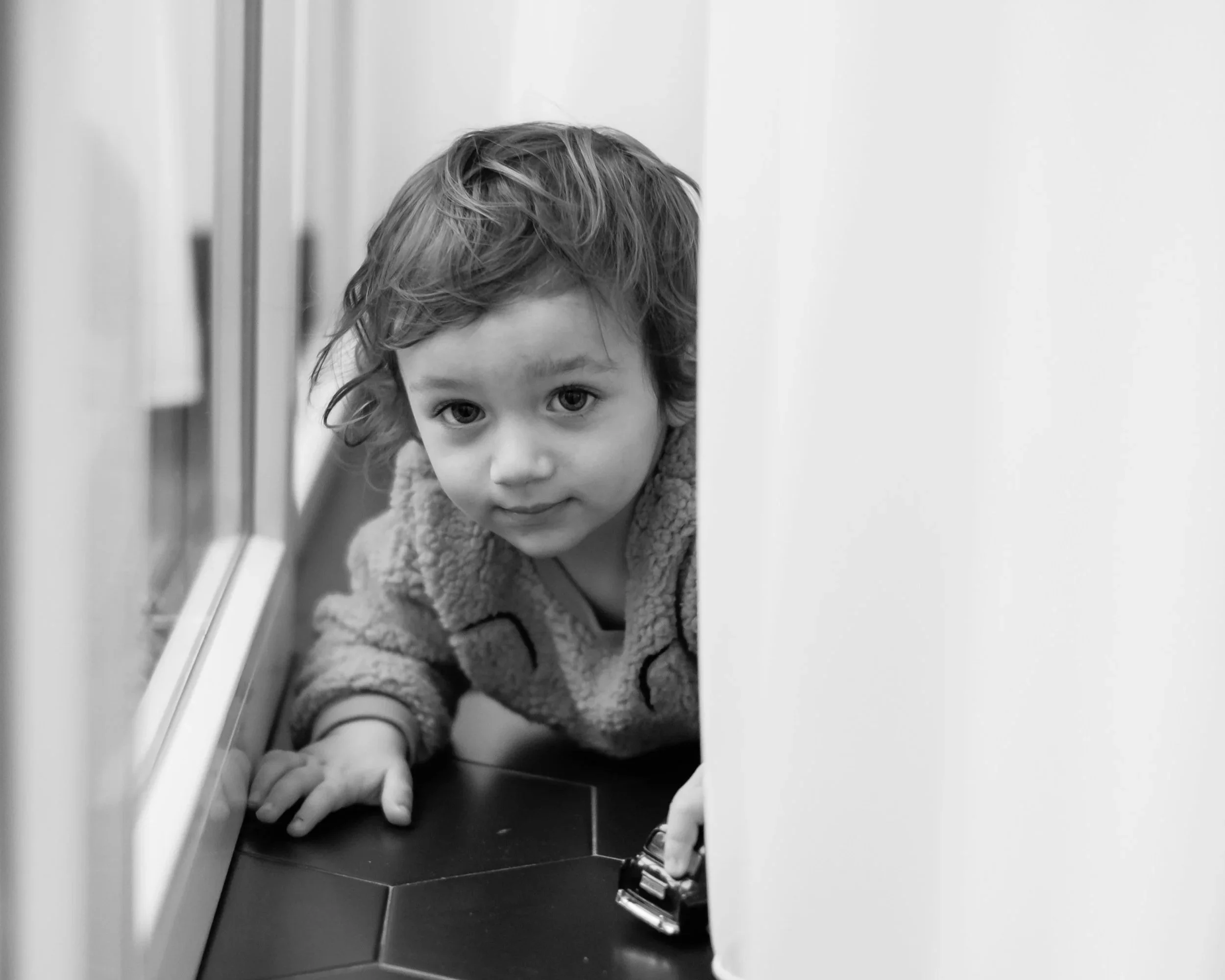 Young child crouching beside a window and looking toward the camera in a black-and-white photograph.
