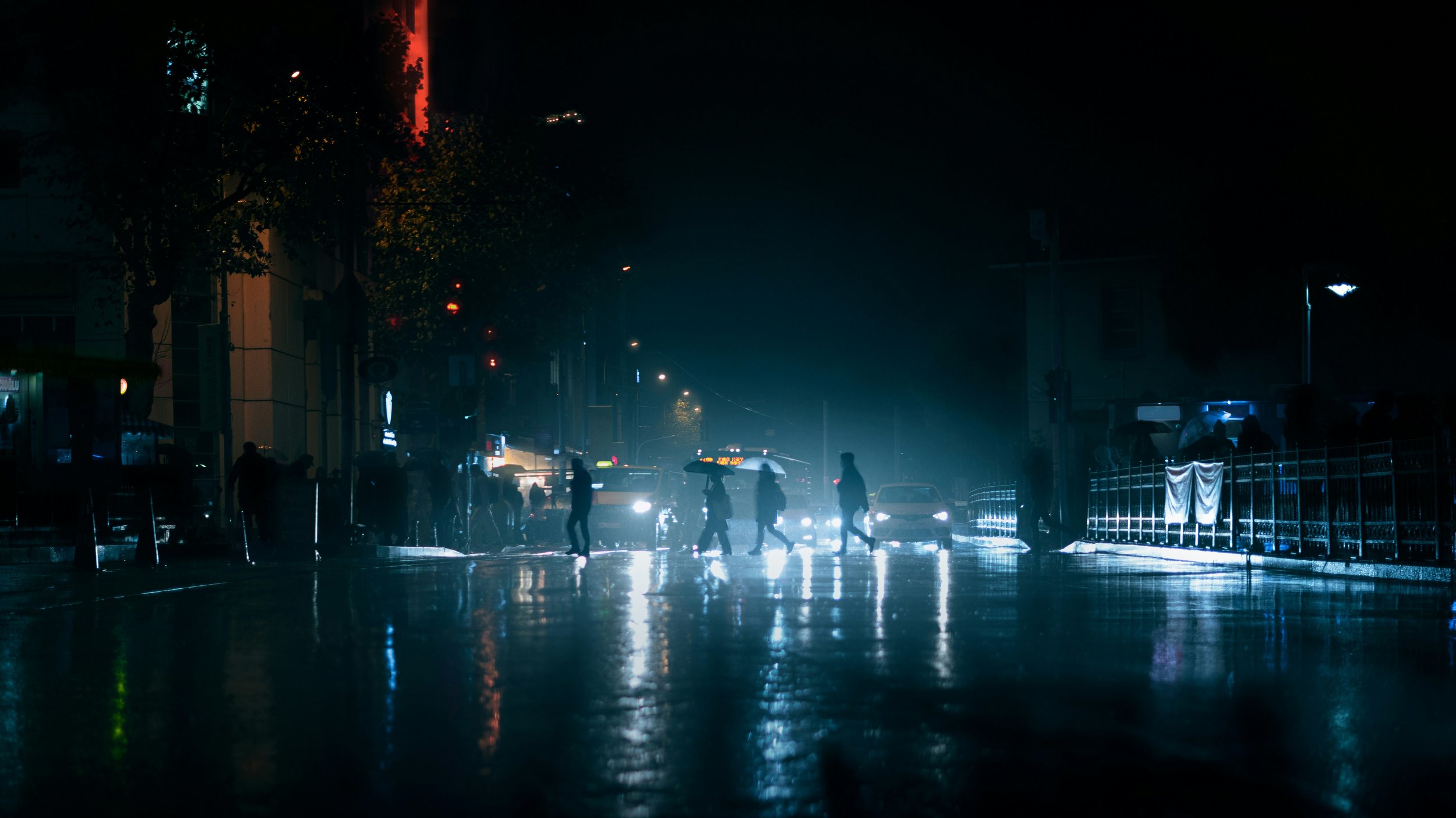 Nighttime city street scene in the rain, with silhouetted pedestrians crossing a reflective wet roadway in front of glowing headlights and neon-lit buildings.