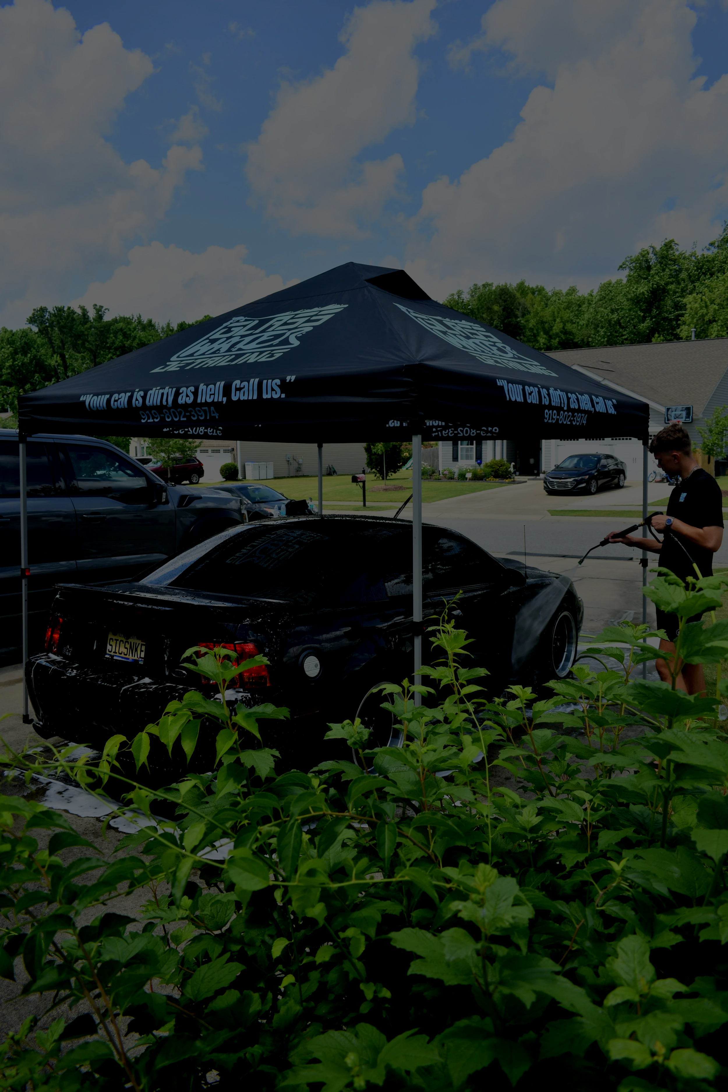Person washing a black car under a black canopy with text, in a residential neighborhood, with bushes in the foreground and other cars parked in the background.