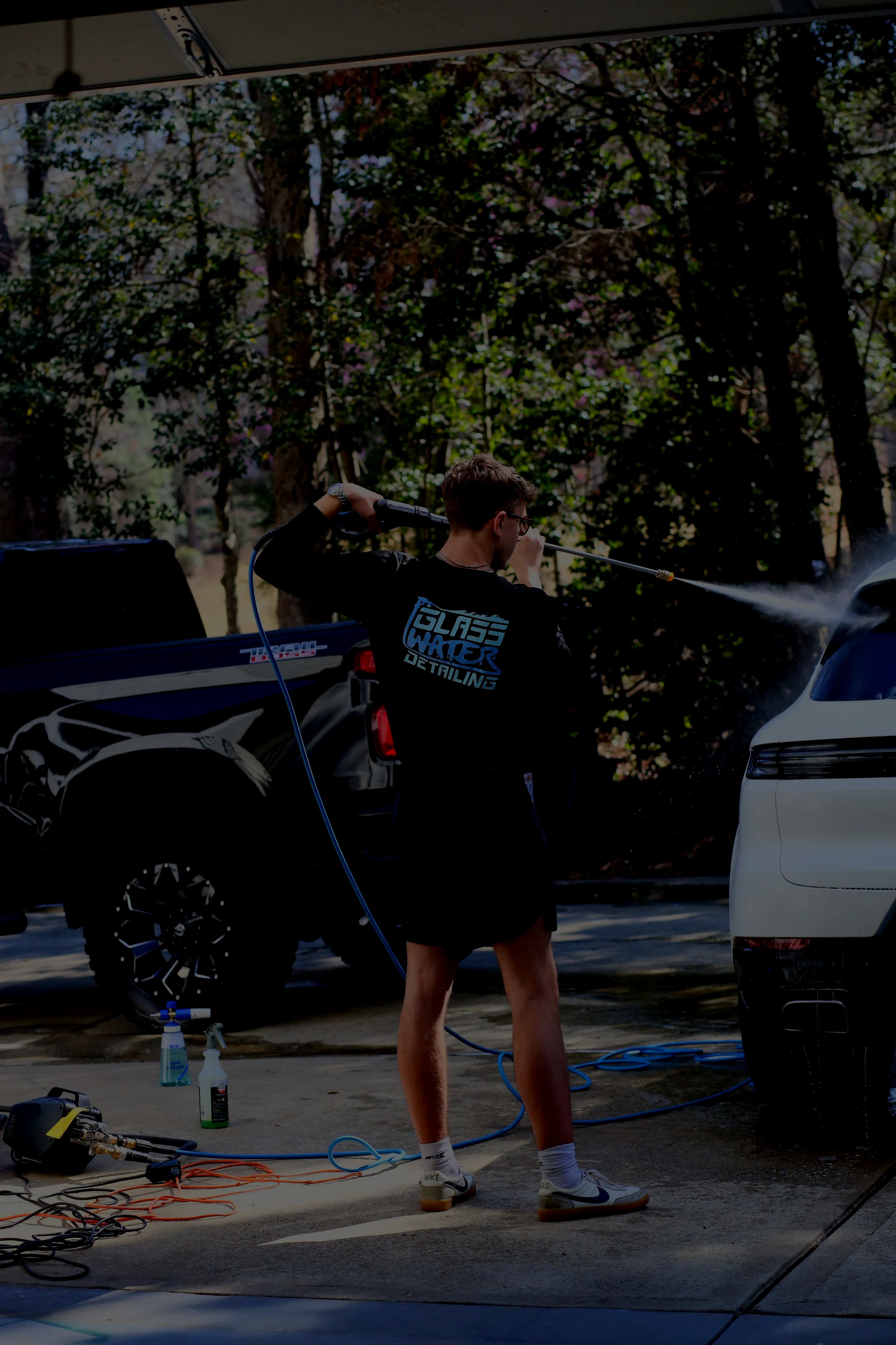 Person washing a white car in a driveway using a pressure washer, with a black pickup truck and trees in the background.