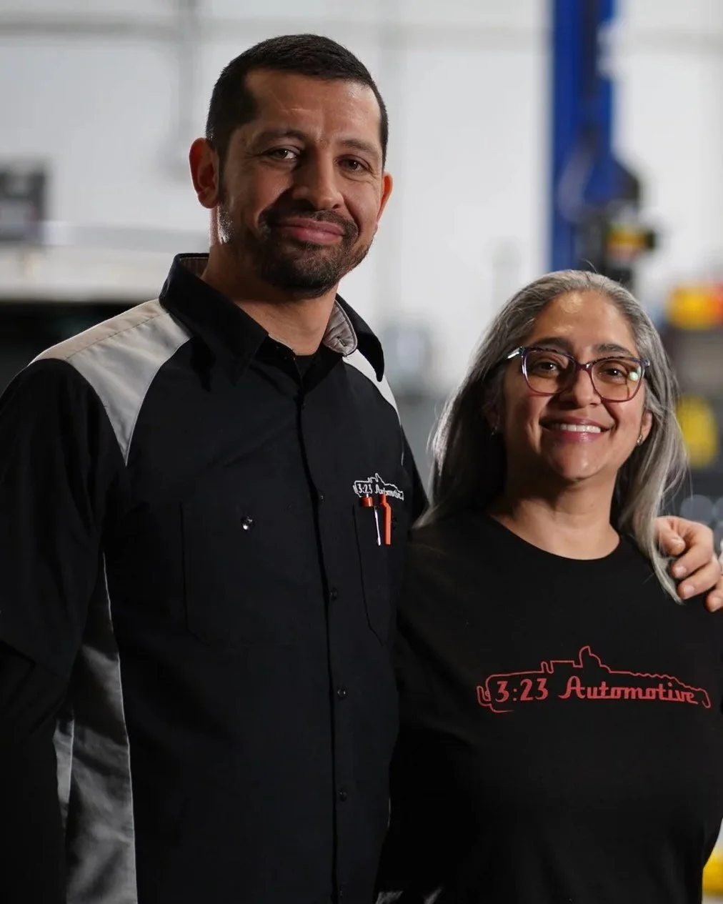 A man with short dark hair and beard, wearing a black shirt with gray accents, standing next to a woman with gray hair, glasses, and a black T-shirt with red text and a car outline, inside a workshop or garage.