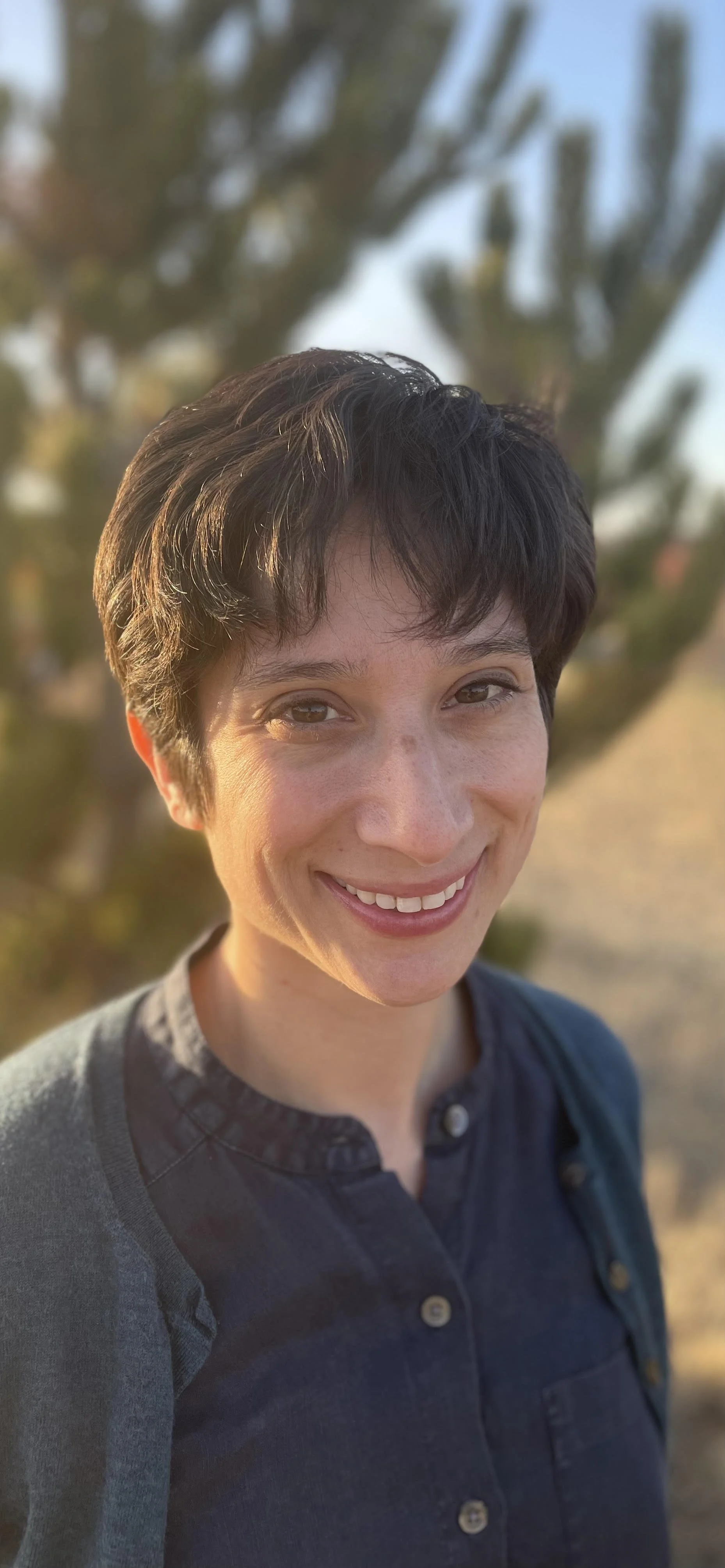 Headshot of Sophia olive skin with short brown hair with an evergreen tree in the background. Grief therapy Longmont, CO
