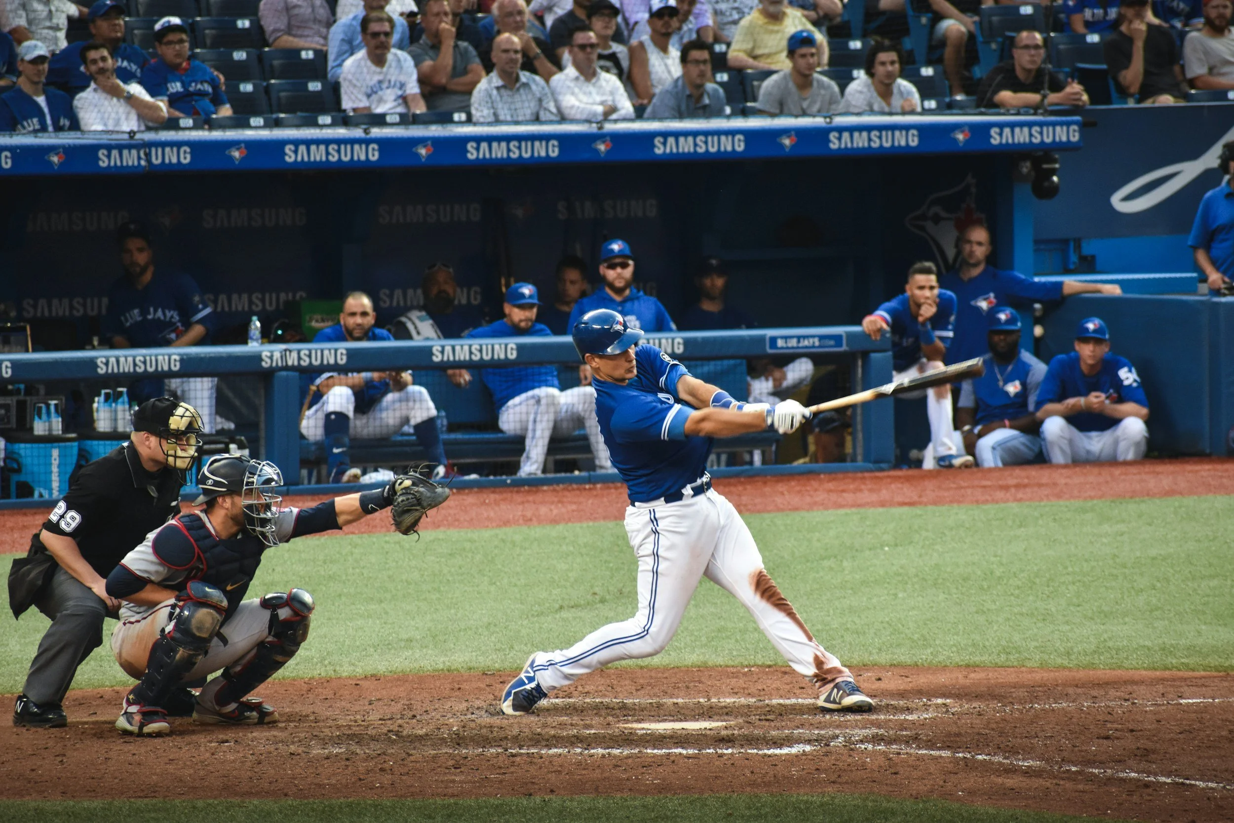 A baseball player in a blue uniform hits a pitched ball at the plate while a catcher and umpire observe. Several spectators sit in the background watching the game.