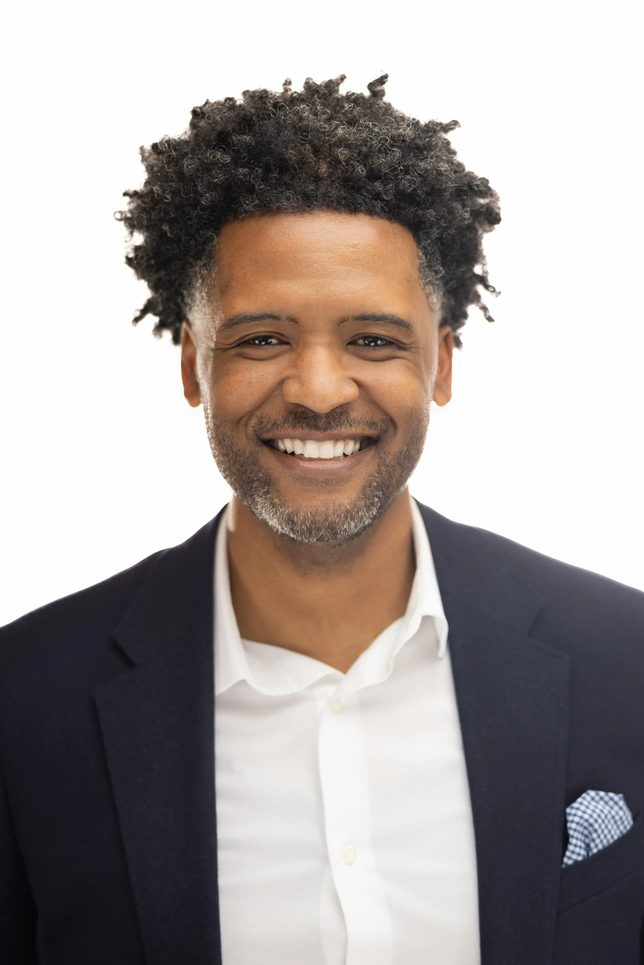 Smiling man with dark curly hair, wearing a white shirt and dark blazer, against a plain white background.