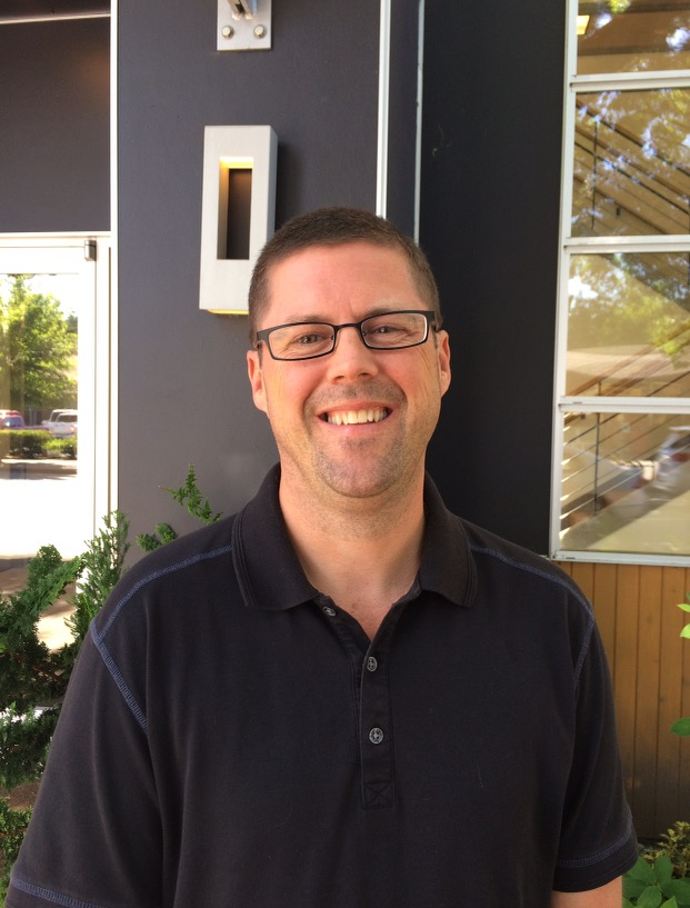 Smiling man with short brown hair and glasses wearing a black collared shirt, standing outdoors near a building with large windows and greenery.