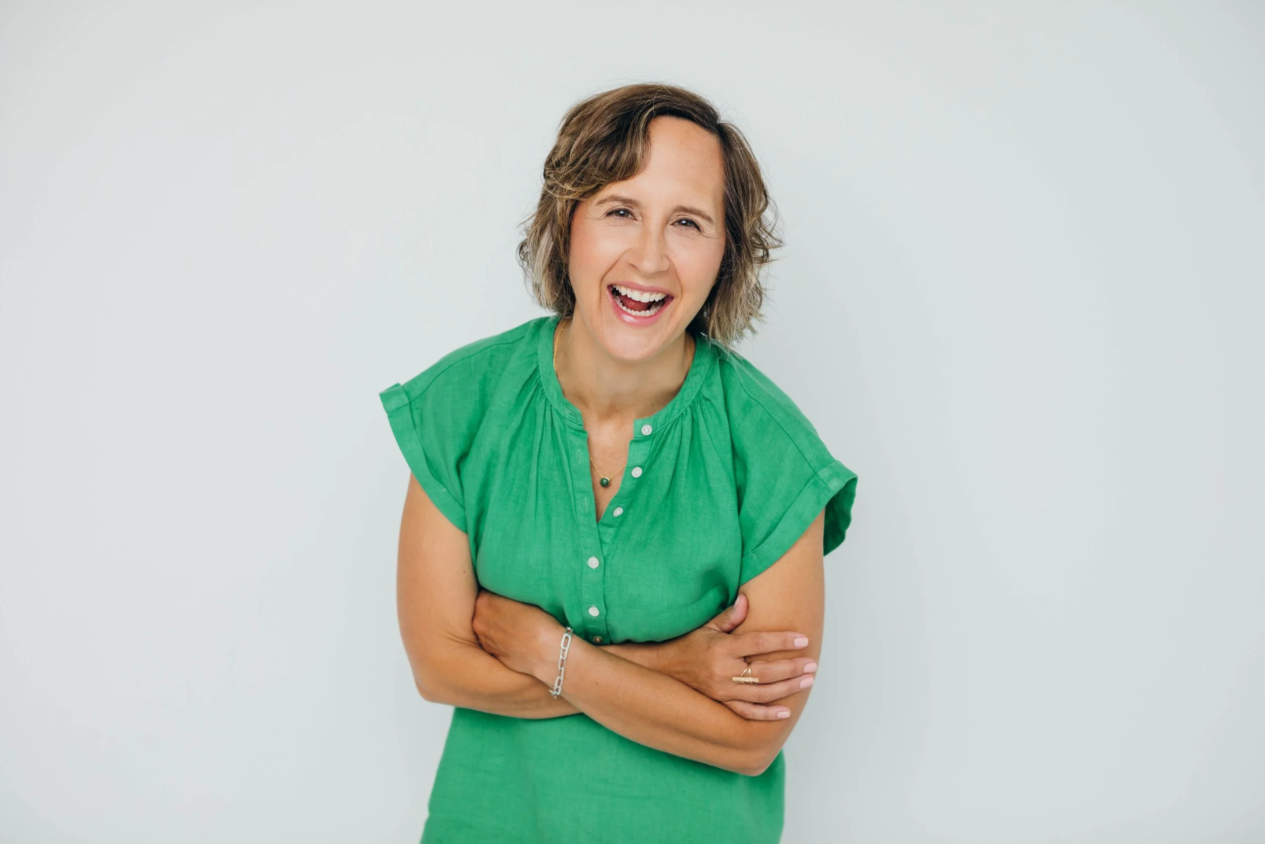 A woman with short, curly brown hair wearing a bright green dress, smiling with her arms crossed in front of her against a plain white background.