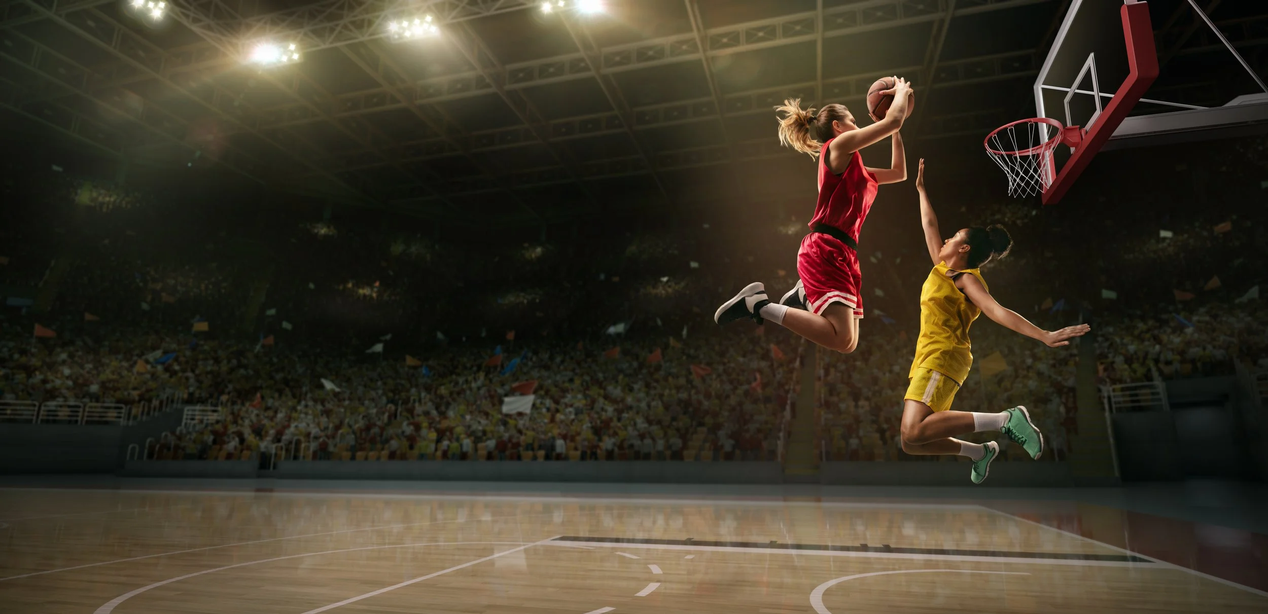 Two female basketball players jumping for the ball near the hoop in an indoor basketball court filled with spectators.