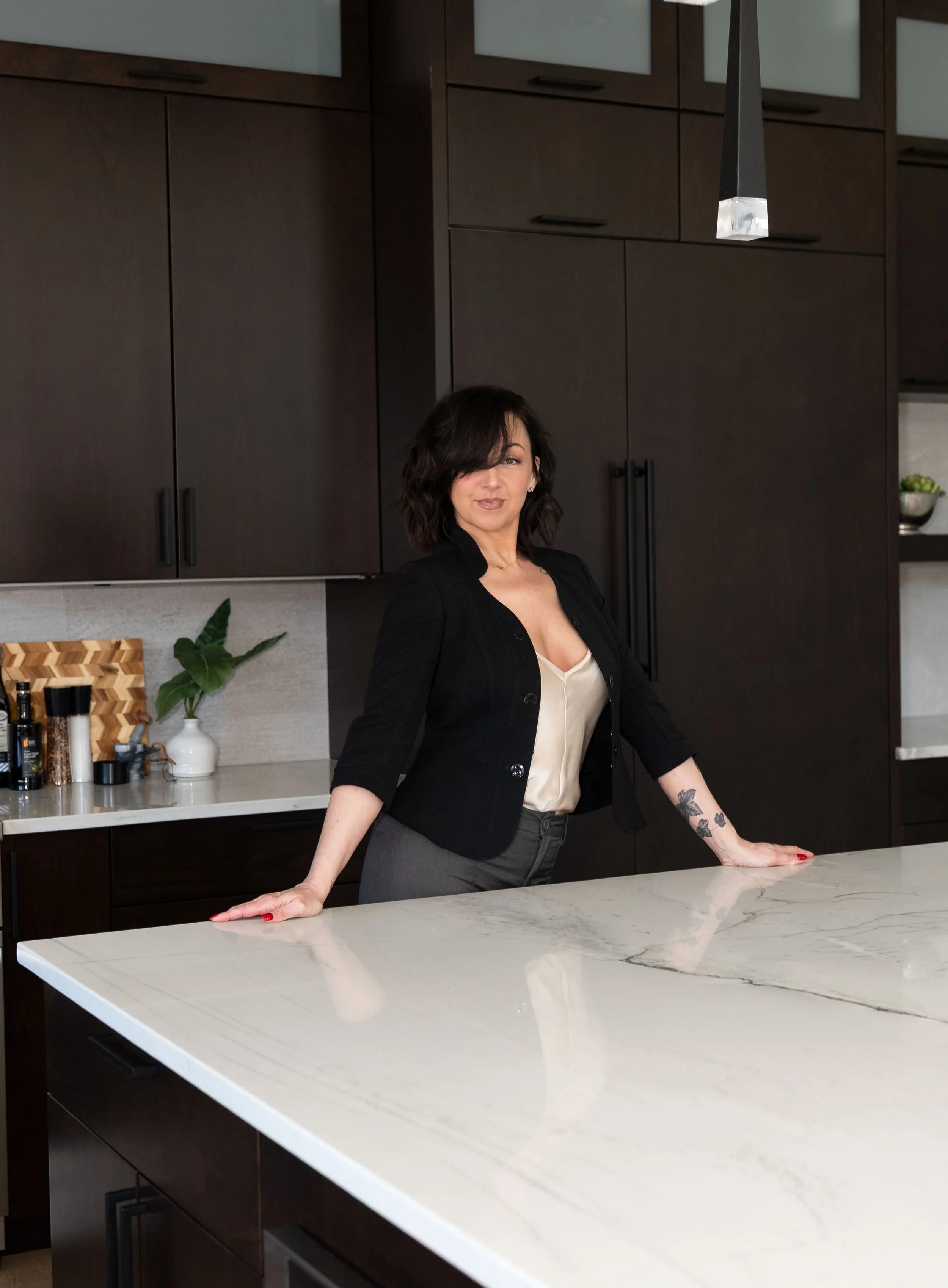 A woman with dark hair and tattoos on her left arm, wearing a black blazer and beige top, leaning on a white marble kitchen countertop with dark wood cabinets in the background.