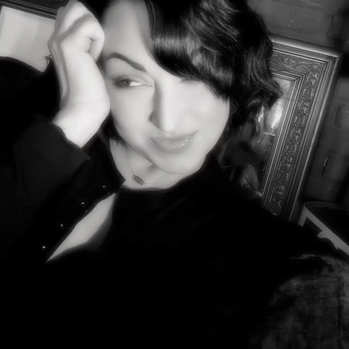 Black and white close-up photo of a woman with short curly hair, smiling and winking, touching her face with one hand, wearing a dark top and a necklace.
