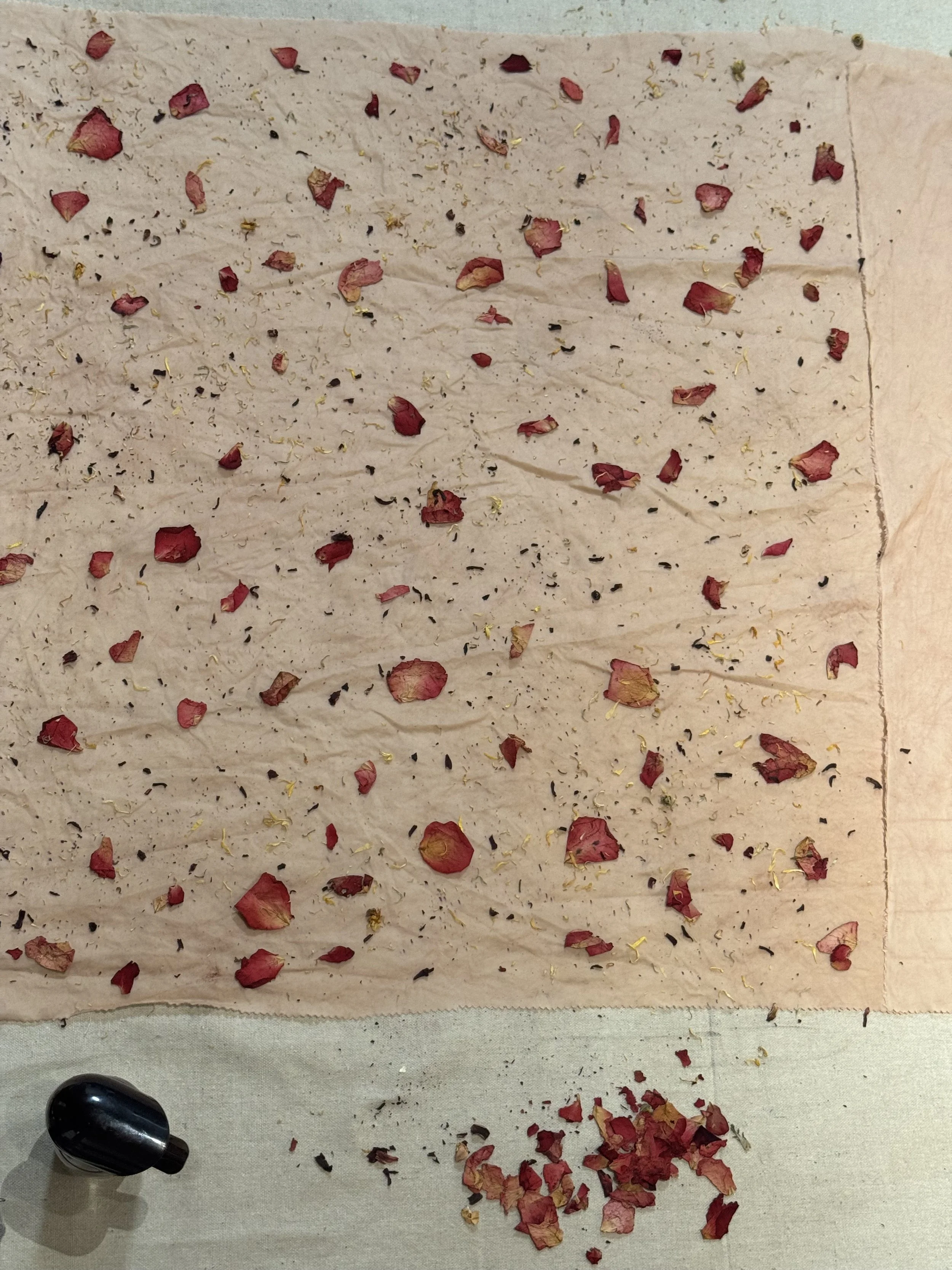 A white sheet of paper with dried flower petals and small black specks scattered across it, placed on a work surface with a black tool near the bottom left corner.