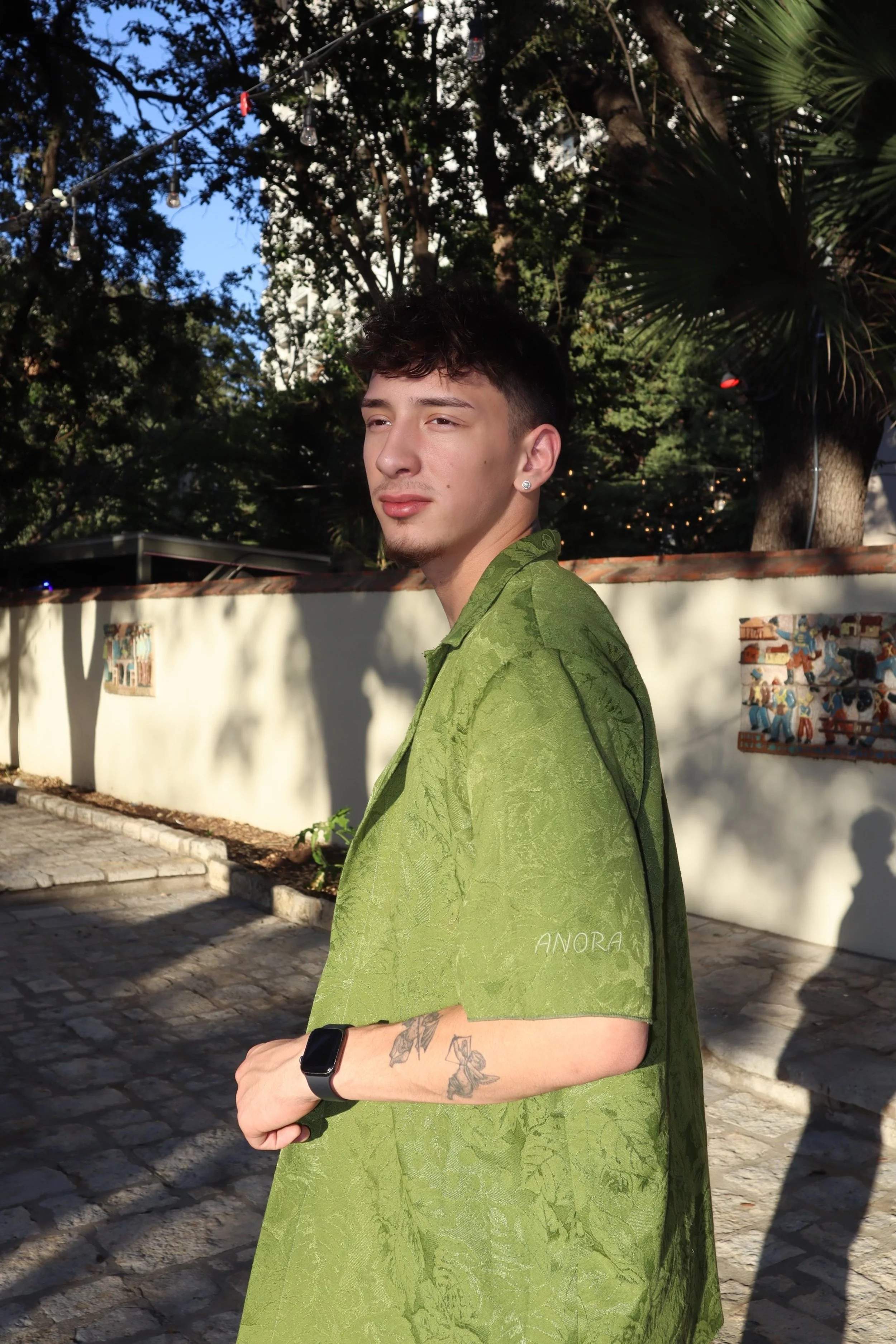 A young man with short dark hair, wearing a green patterned shirt, standing outdoors on a cobblestone street with a white wall and trees in the background. The sunlight is casting shadows, and he has tattoos on his left forearm and is wearing a black smartwatch.