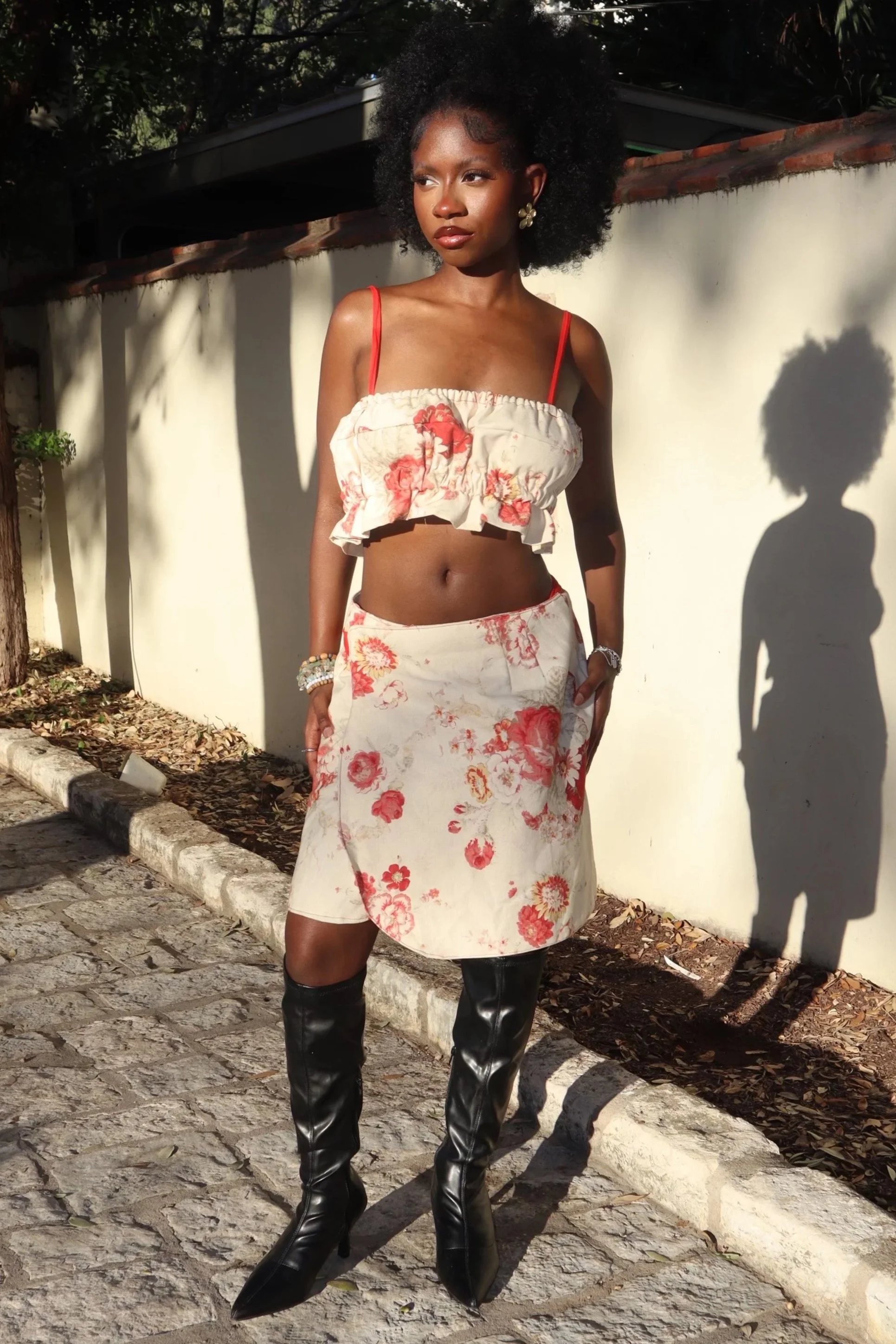 A woman with curly afro hair wearing a floral crop top, matching floral skirt, and black knee-high boots, standing outdoors on cobblestone with sunlight casting shadows.