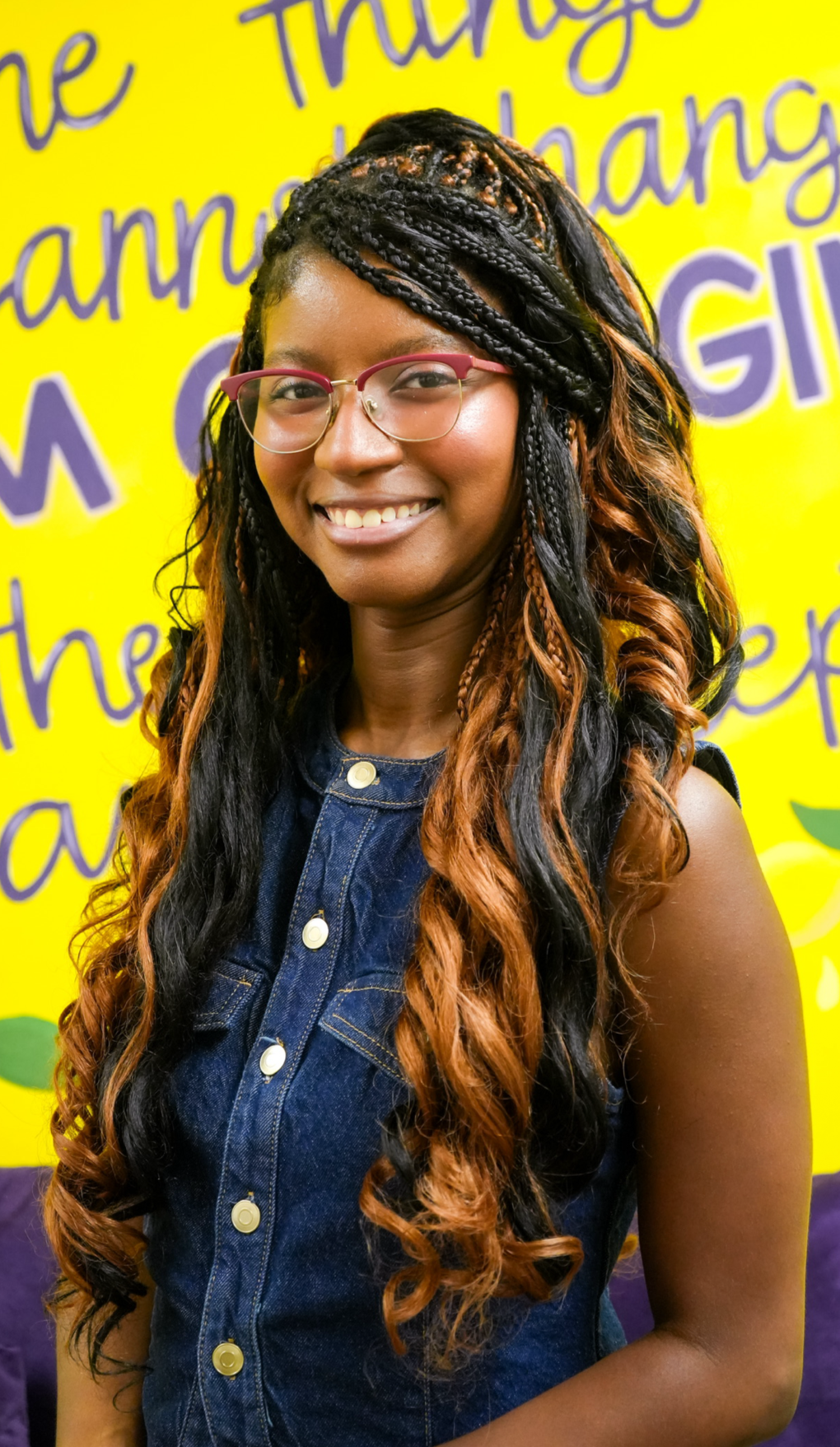 Young woman with long, wavy, black and brown hair, wearing glasses and a sleeveless denim top, smiling against a bright yellow background with purple and green writing and graphics.