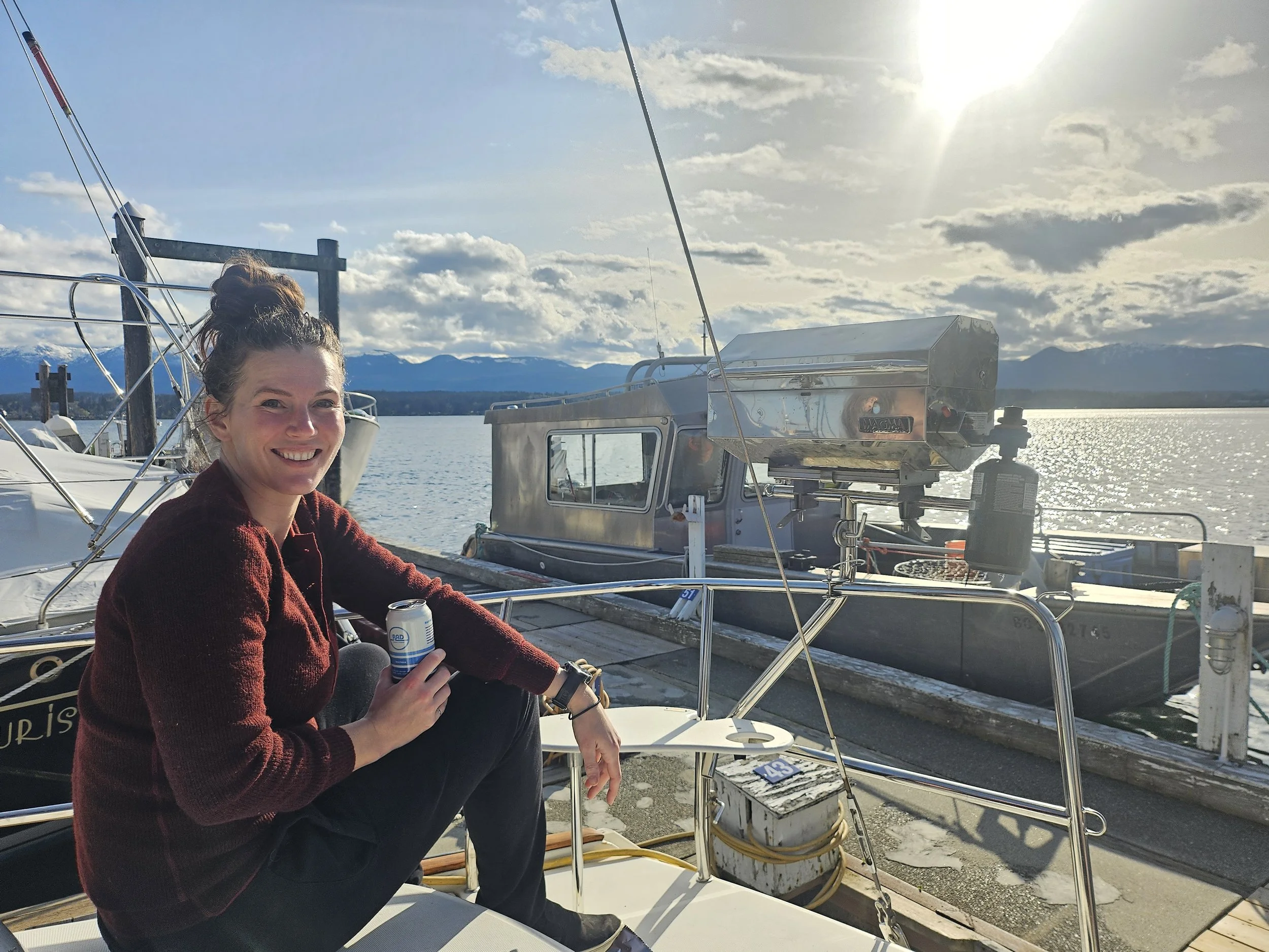 A woman sitting on a boat holding a can of beer, smiling, with a marina and mountains in the background and the sun shining in the sky.
