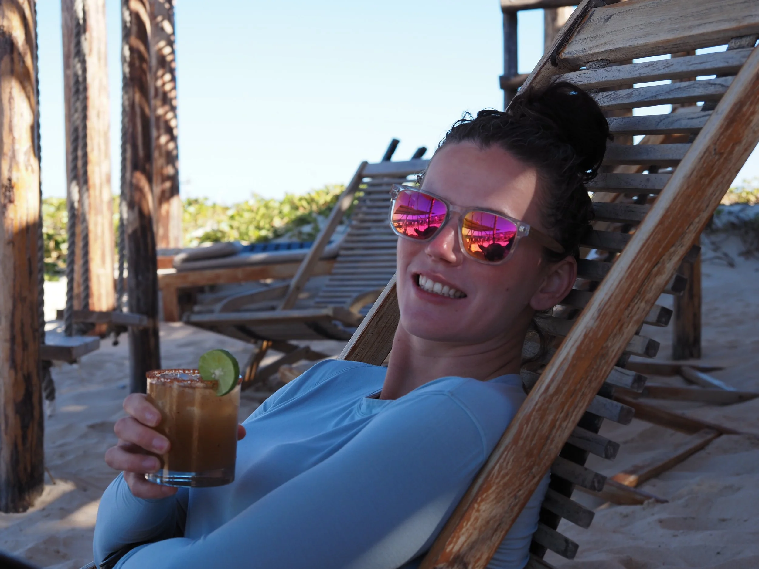 Woman relaxing on a wooden lounge chair at the beach, wearing sunglasses and holding a drink with a lime garnish.