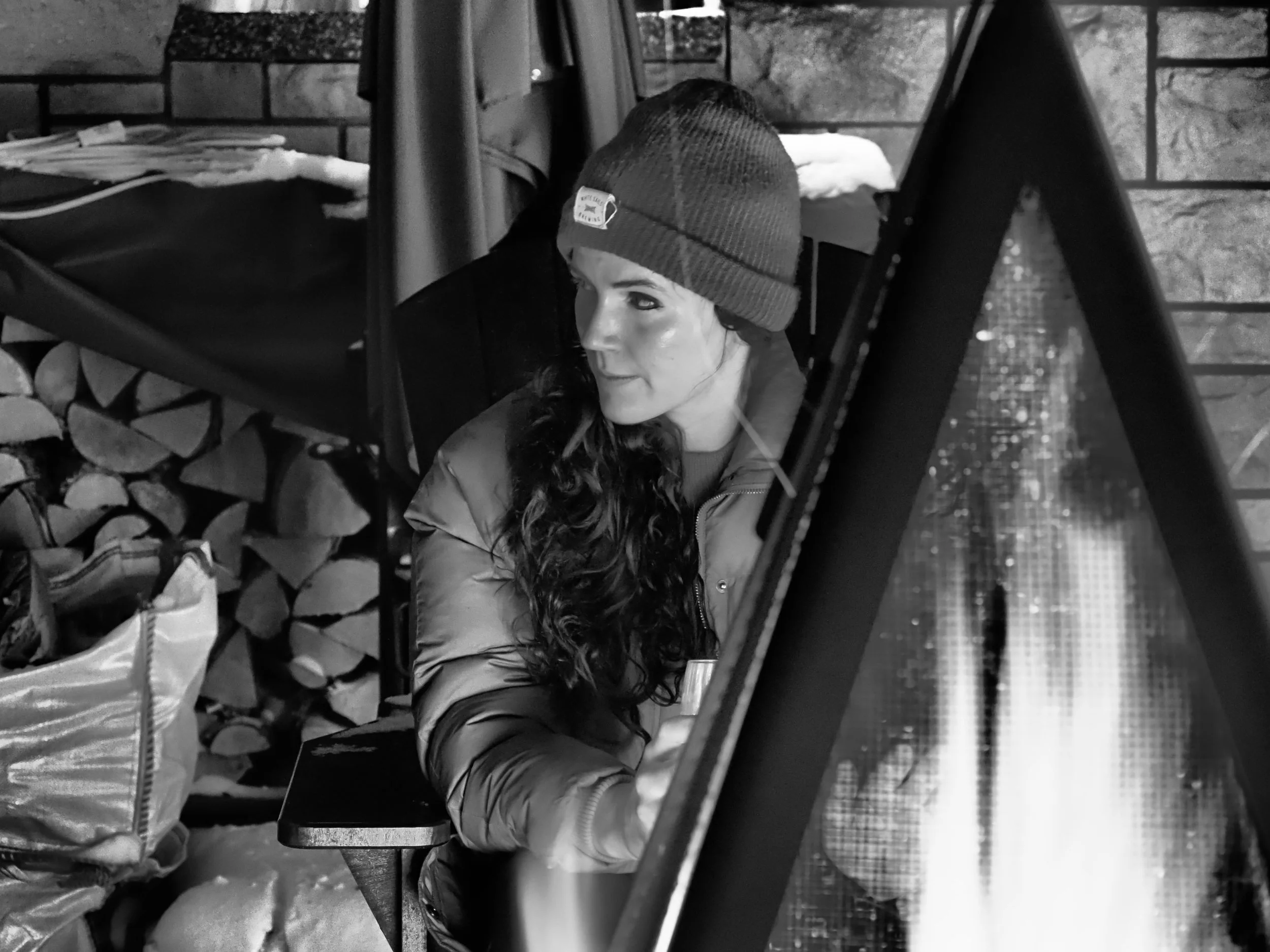 A woman with long curly hair wearing a beanie and puffy jacket, sitting outdoors near a fireplace with stacked firewood behind her.