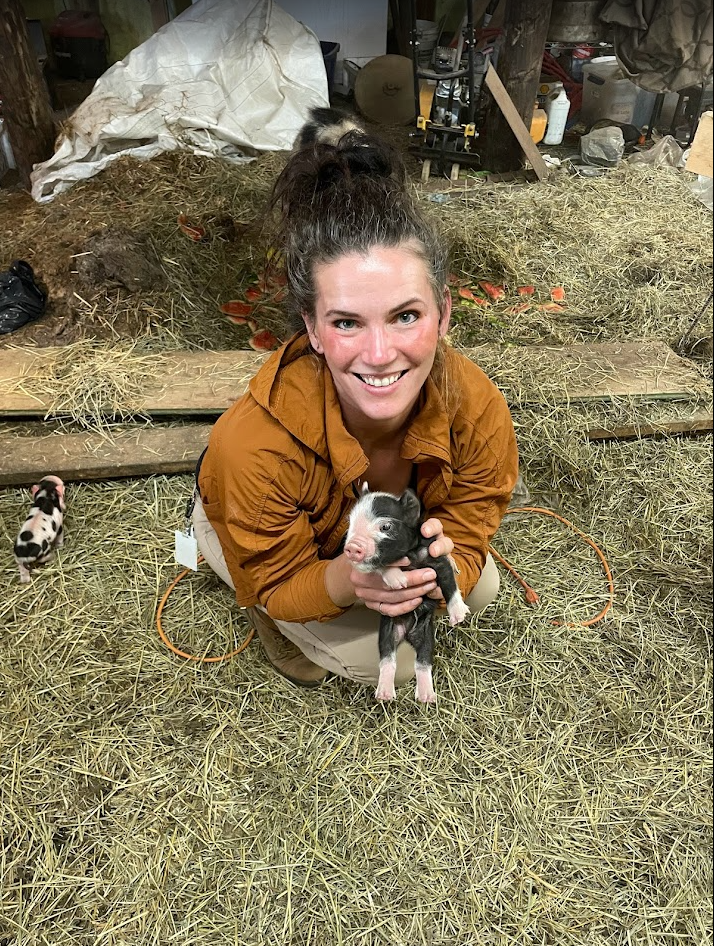 A smiling woman with dark hair tied back, wearing a brown jacket, is holding a tiny black and white piglet in a barn or farm setting. The piglet has a pink snout and ears. The background shows hay, some gardening or farm tools, and supplies.