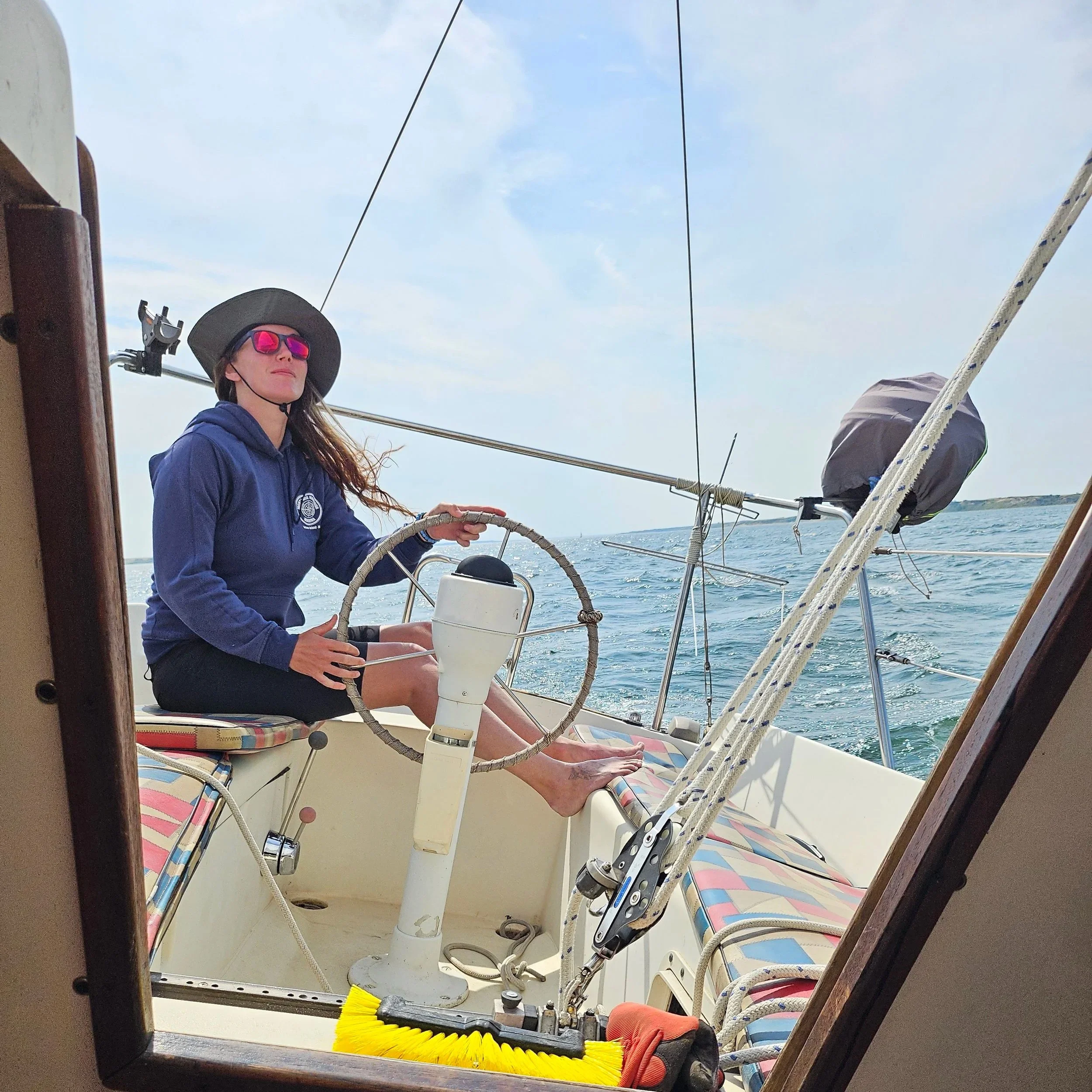A woman wearing a hat, sunglasses, and a blue hoodie sailing a boat on the water.