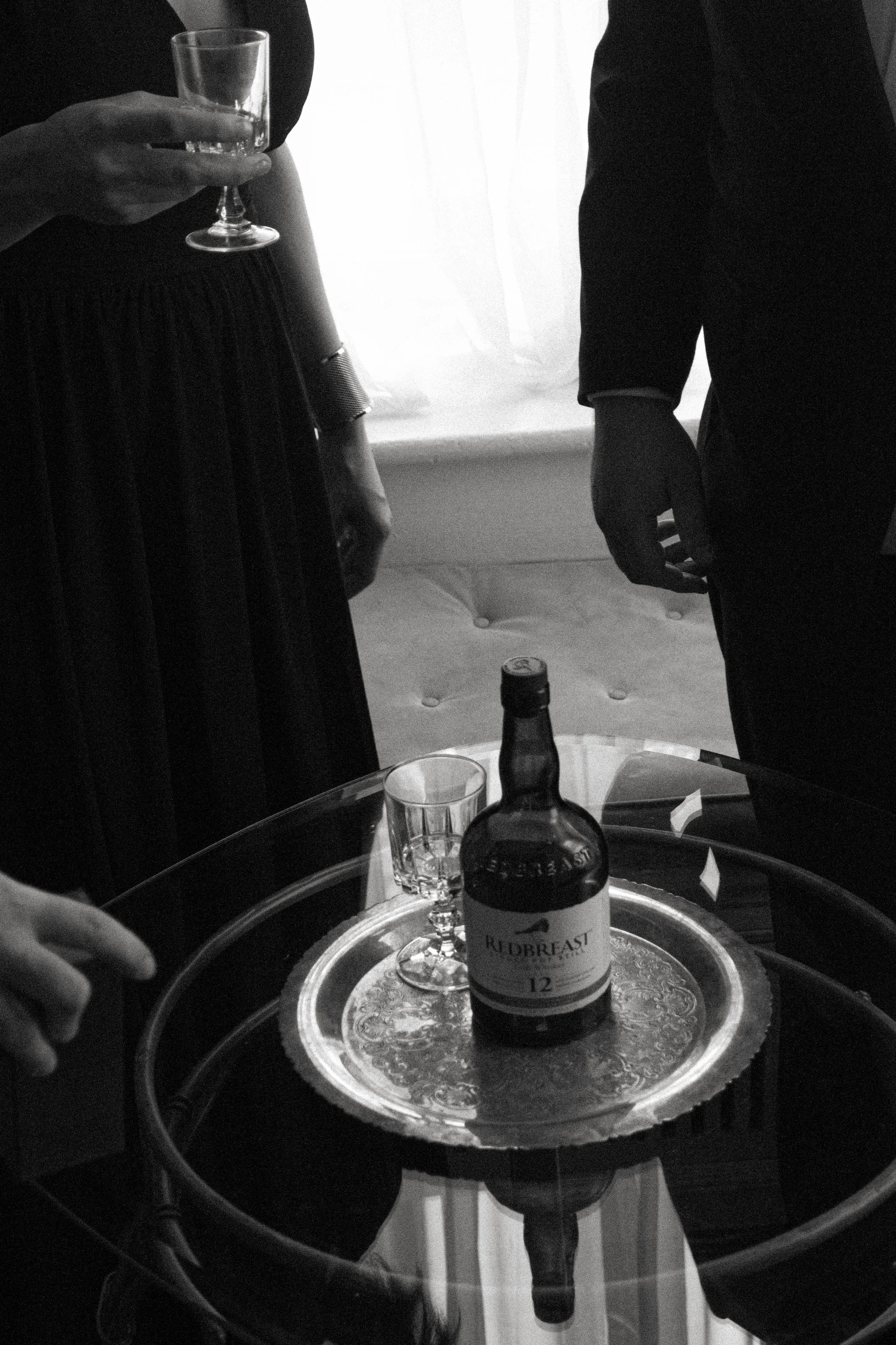 Verre à vin vide, bouteille de whisky, verre vide, et une boîte à liqueur sur une table en verre avec un plateau argenté, dans une pièce sombre avec un éclairage naturel.