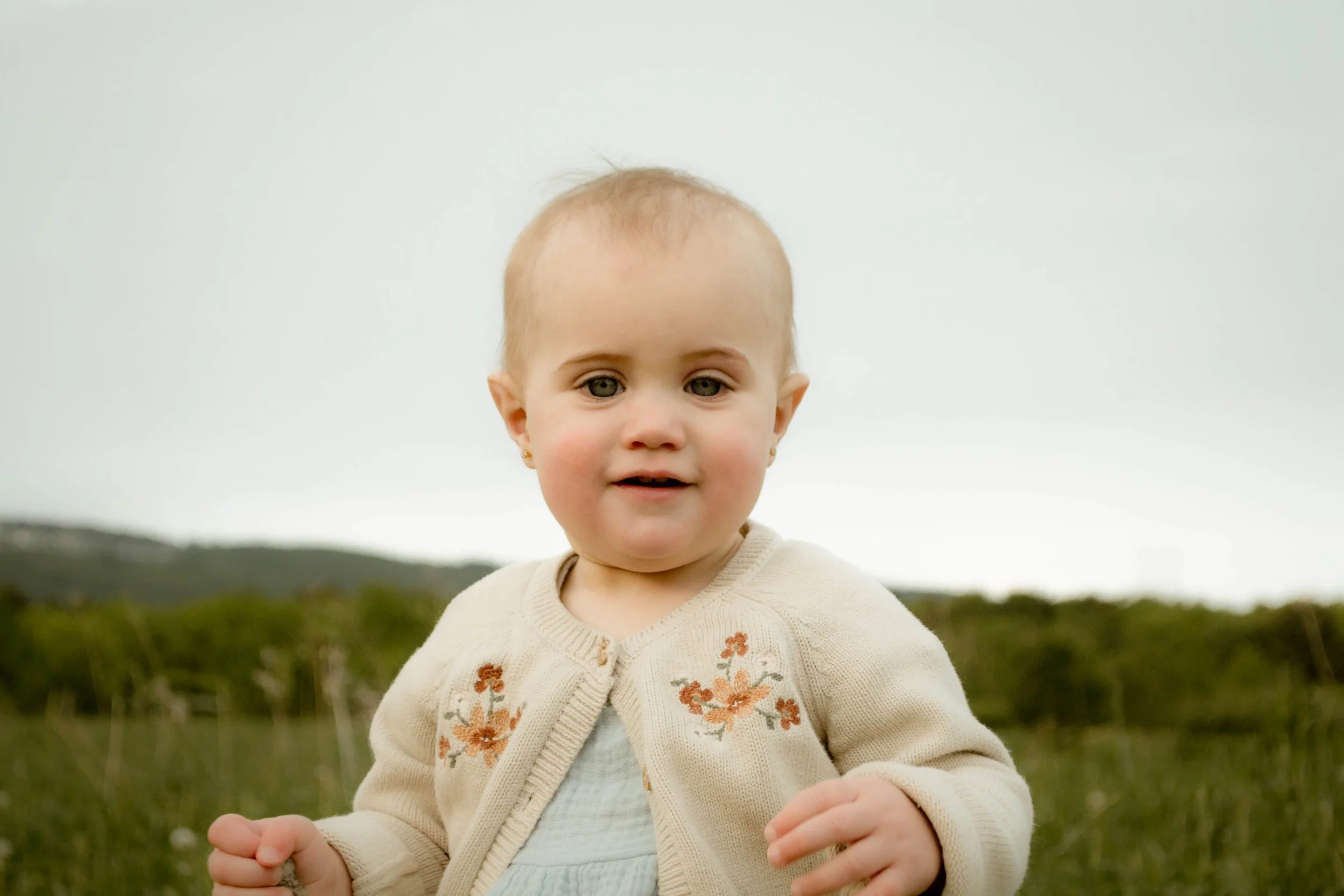 Jeune enfant avec des cheveux clairs et des yeux verts, portant un cardigan beige avec des fleurs d'orange, se tenant en plein air dans un paysage naturel avec une végétation verte et un ciel gris.