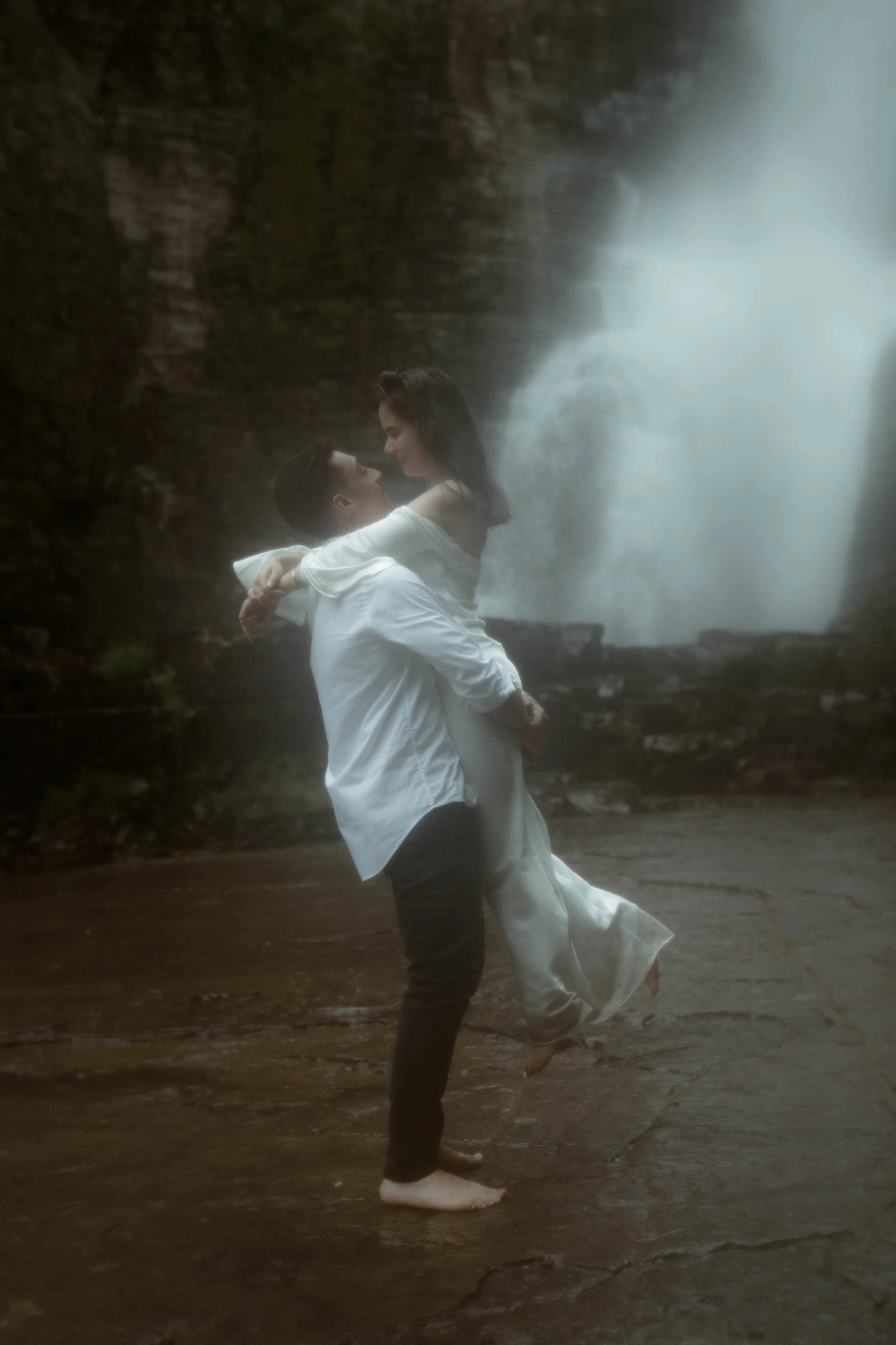 Un couple dansant au bord d'une cascade sous une pluie fine, avec des vêtements blancs et noirs, en position de danse romantique.