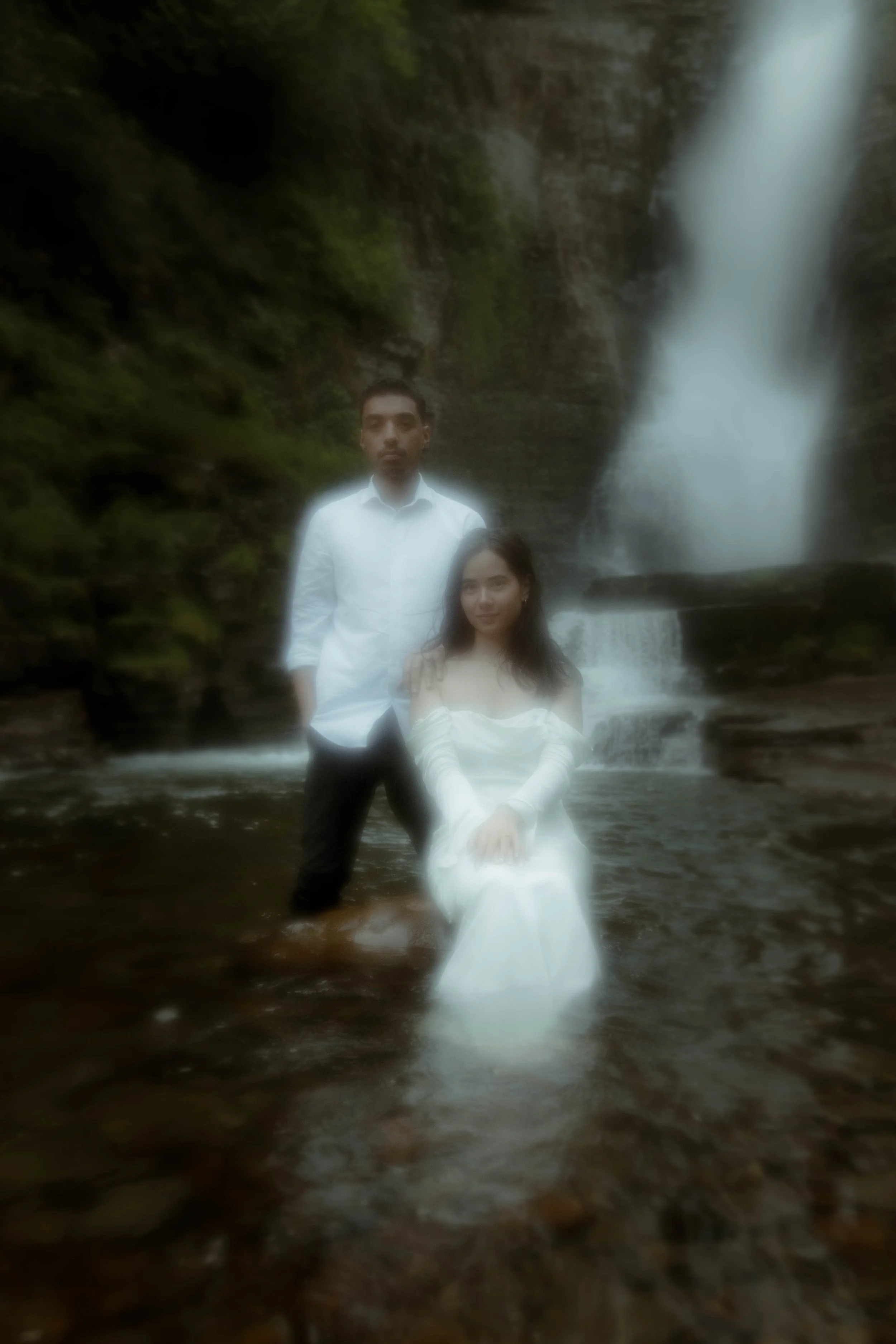 Un homme et une femme posant devant une cascade dans un environnement naturel, l'homme debout et la femme assise sur une roche dans l'eau.