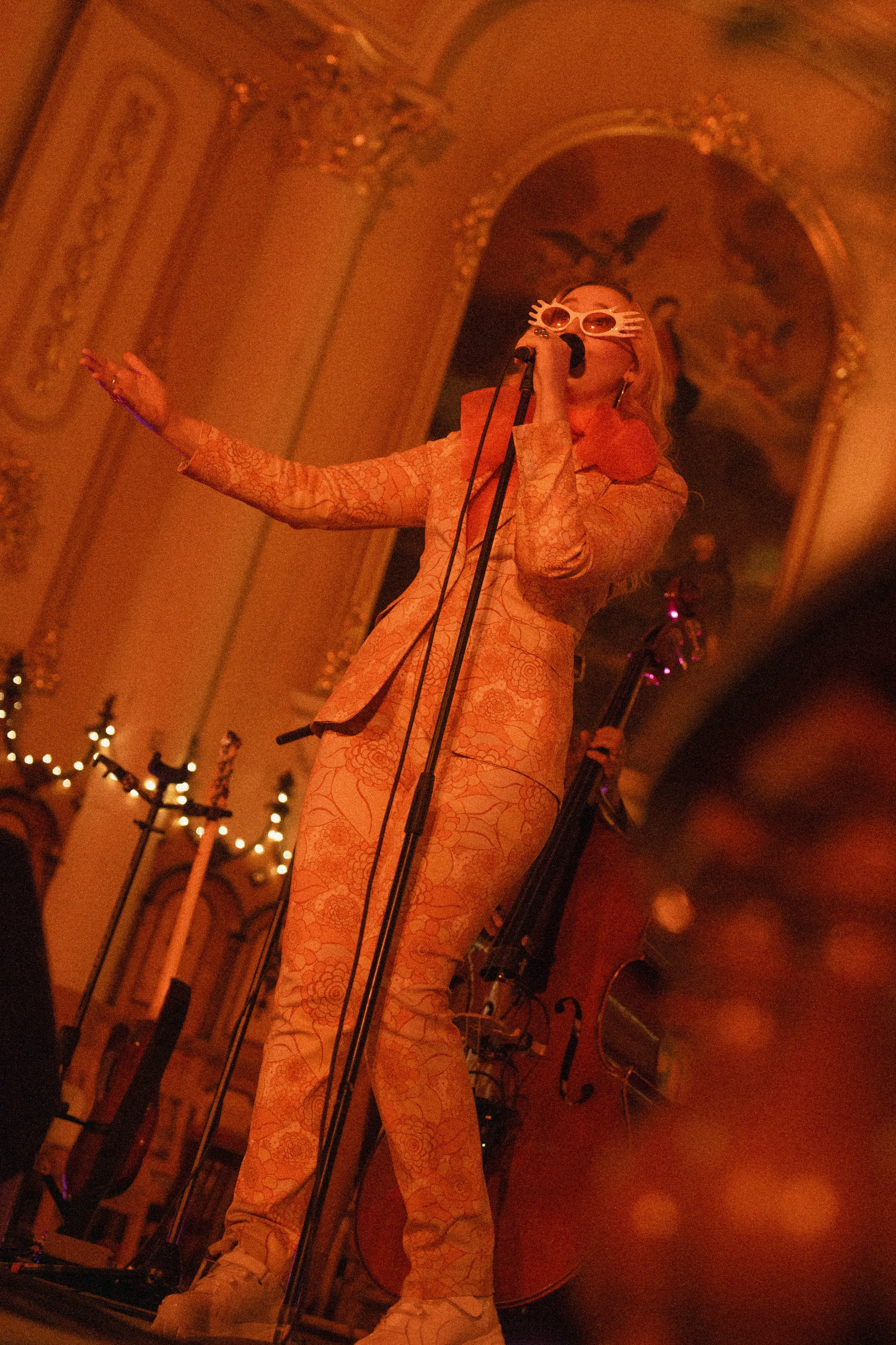 Une femme chante sur scène dans une salle ornée avec des décorations dorées et des lumières chaudes, portant une tenue à motif floral clair et des lunettes de soleil. Elle tient un microphone et semble faire un spectacle ou une performance musicale.