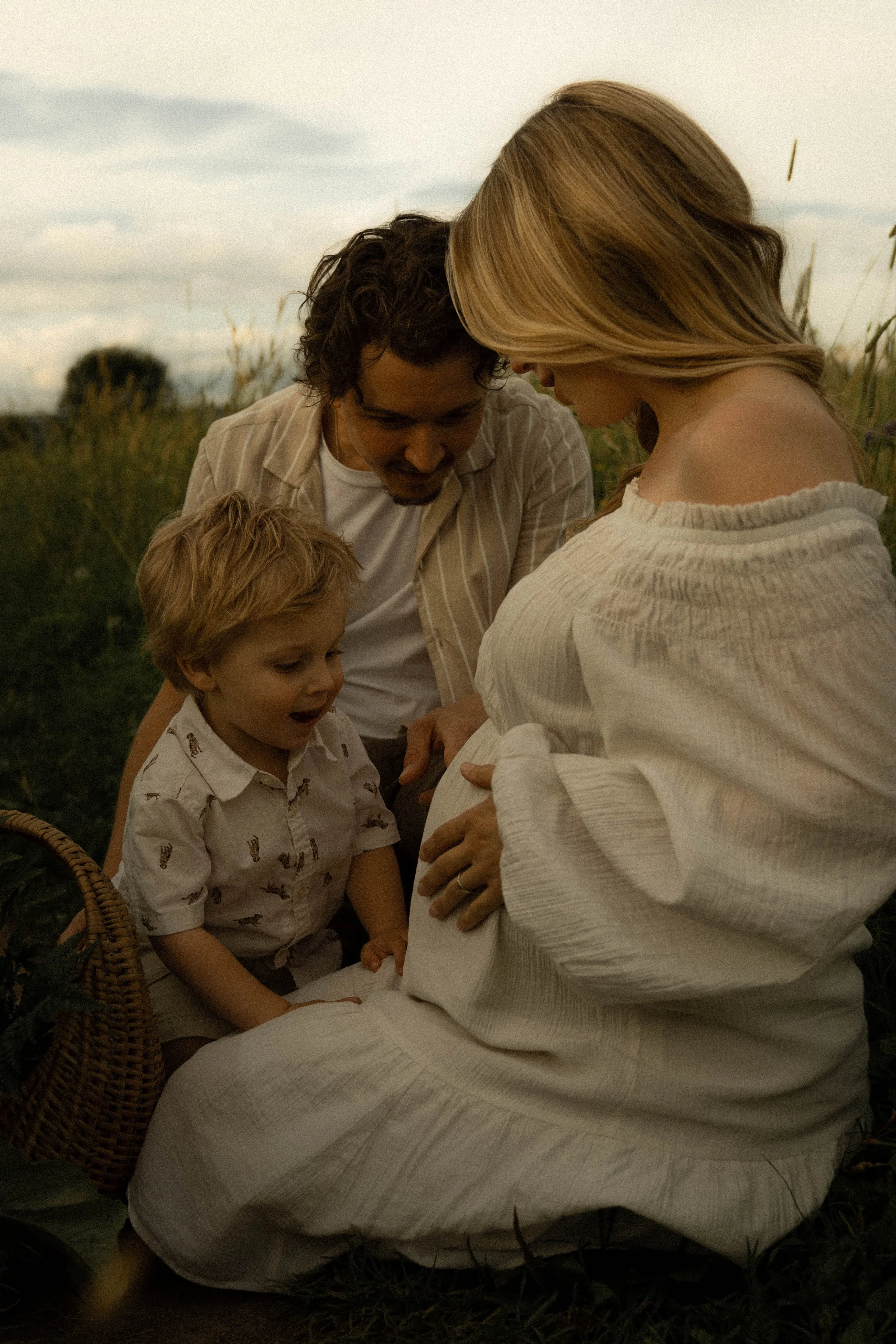 Une femme enceinte assise dans un champ d'herbes, entourée d'un homme et d'un garçon, tous regardant la même direction. Ambiance chaleureuse lors du coucher ou lever du soleil.