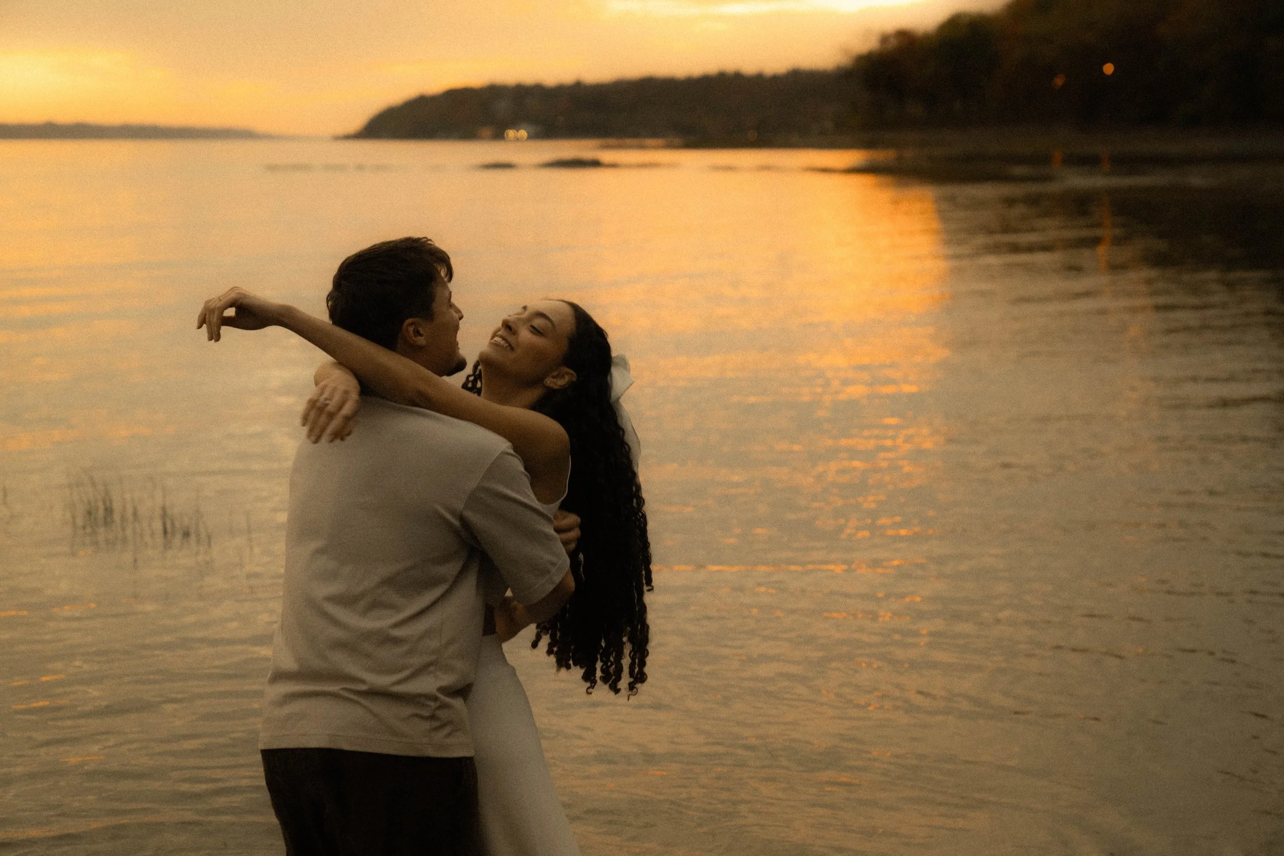 Un couple en train de s'étreindre près de la plage au coucher du soleil, avec des eaux calmes et un paysage de côte en arrière-plan.