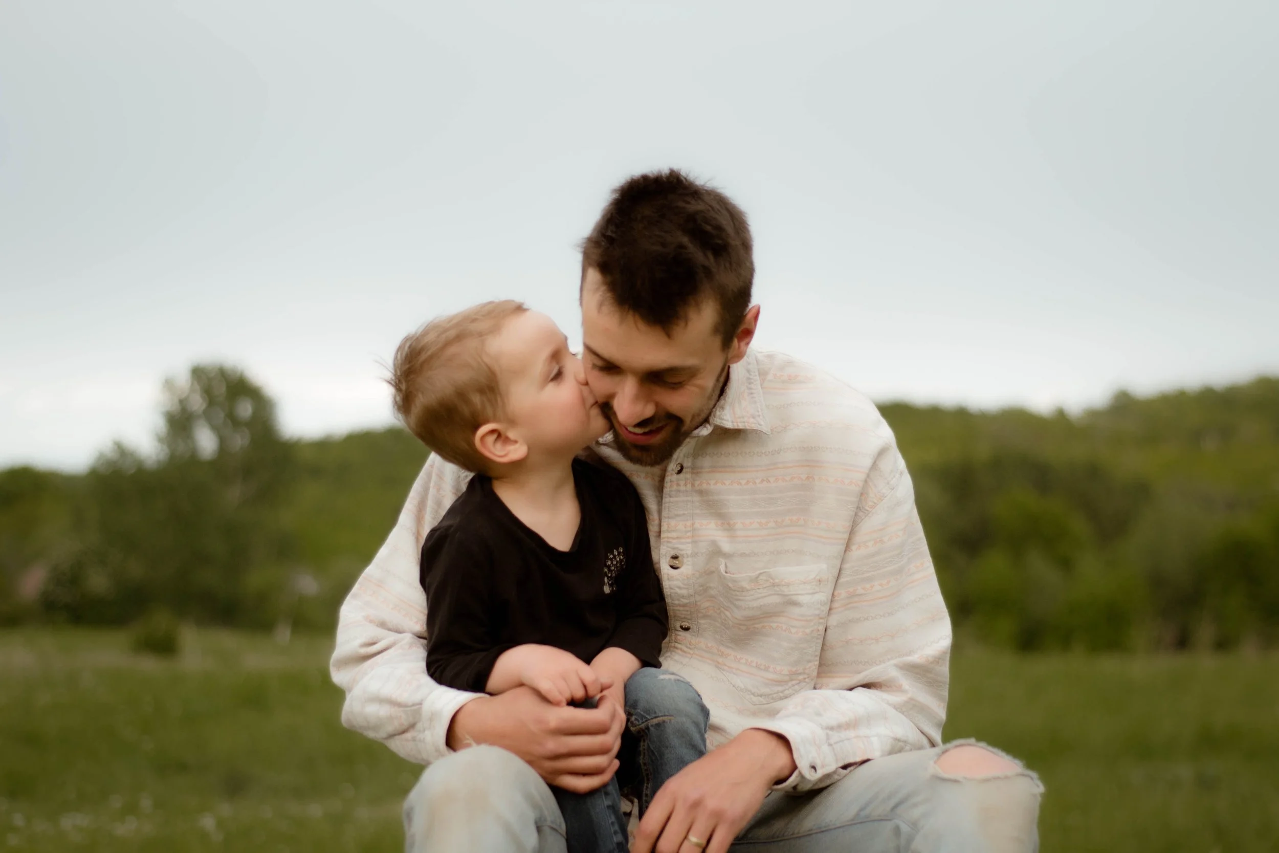 Un jeune garçon donne un bisou affectueux à un homme, probablement son père, dans un parc avec un ciel gris en arrière-plan.