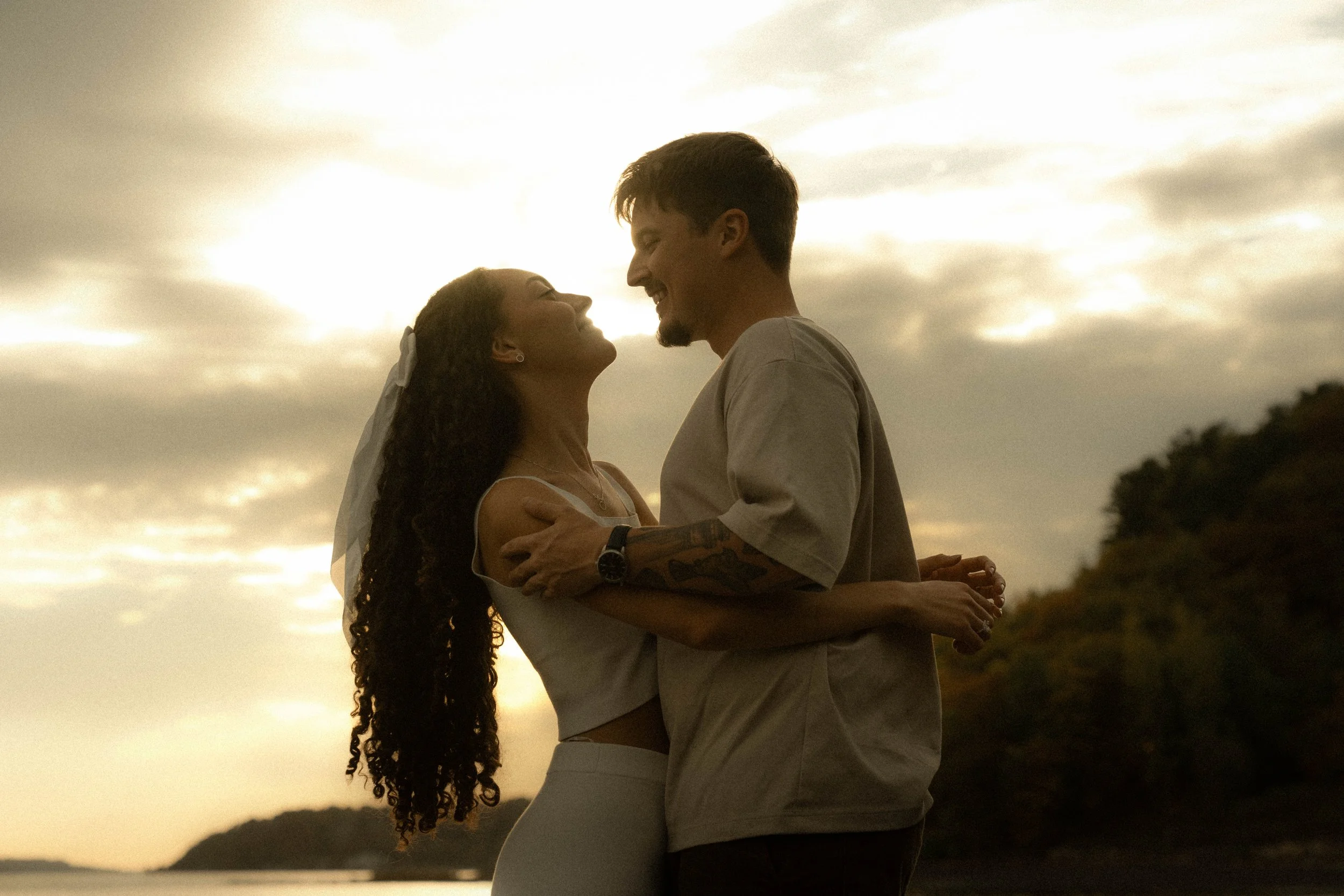 Un couple se regarde et s'embrasse devant un coucher de soleil, avec un ciel nuageux et des collines en arrière-plan.