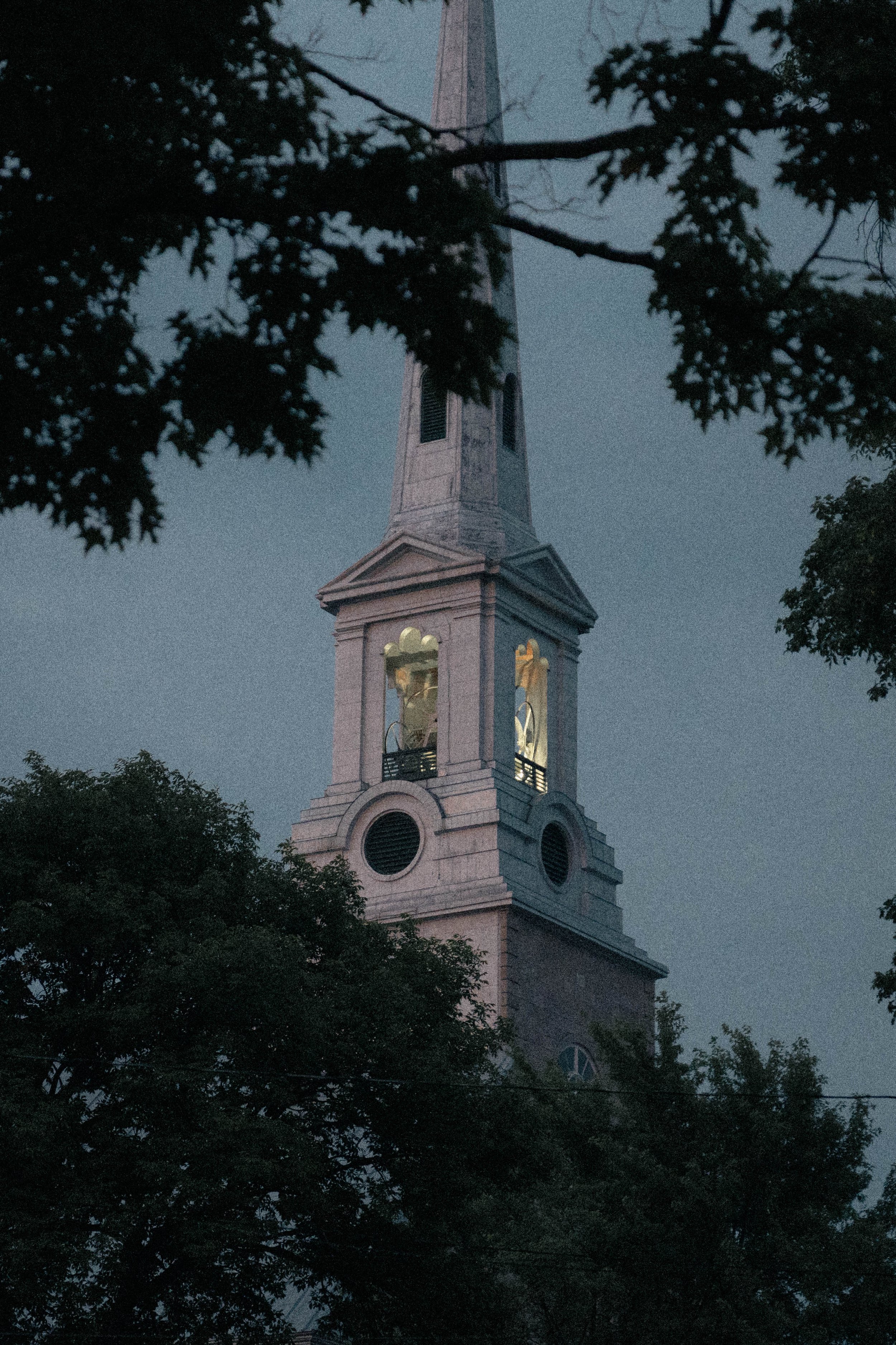 Une tour d'église en pierre blanche vue à travers des branches d'arbres, en fin de soirée ou à l'aube, avec une lumière douce dans le ciel.