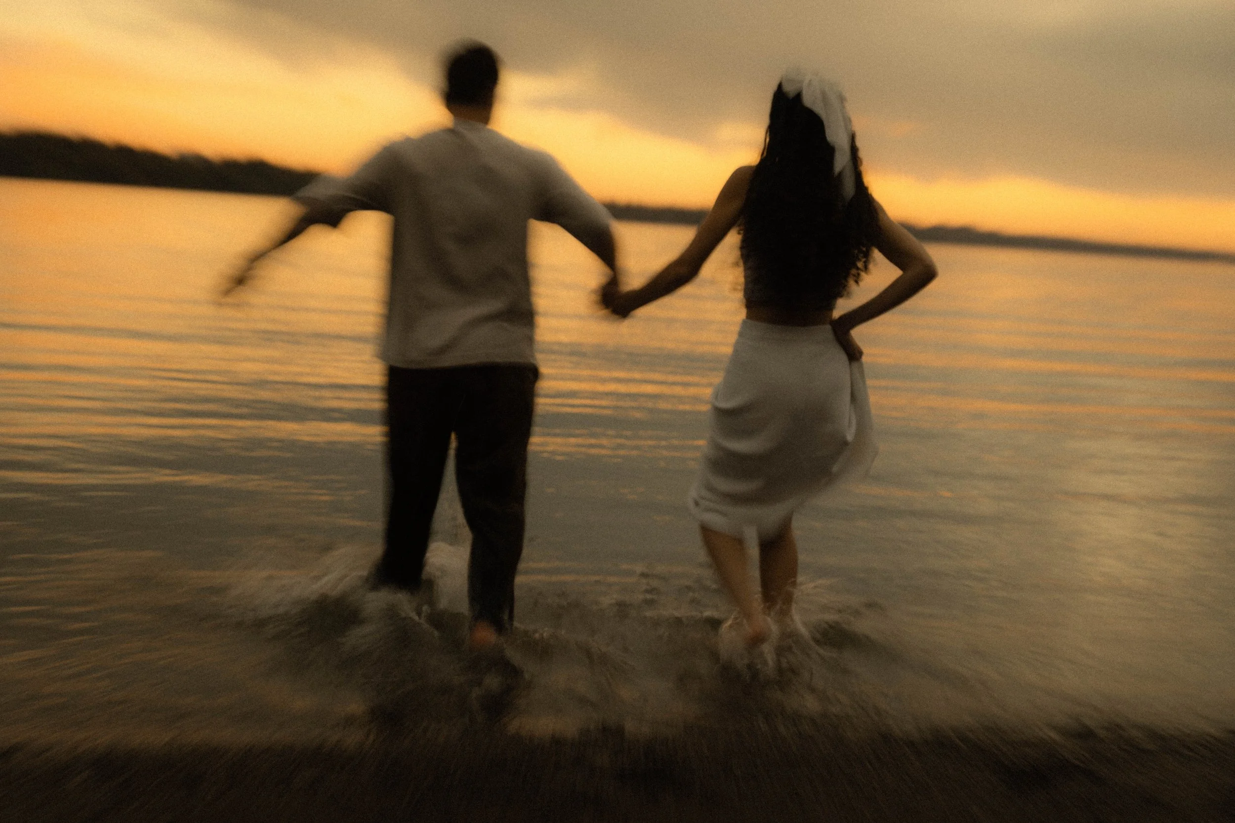 Deux personnes tenant la main, marchant dans l'eau au coucher du soleil sur la plage.