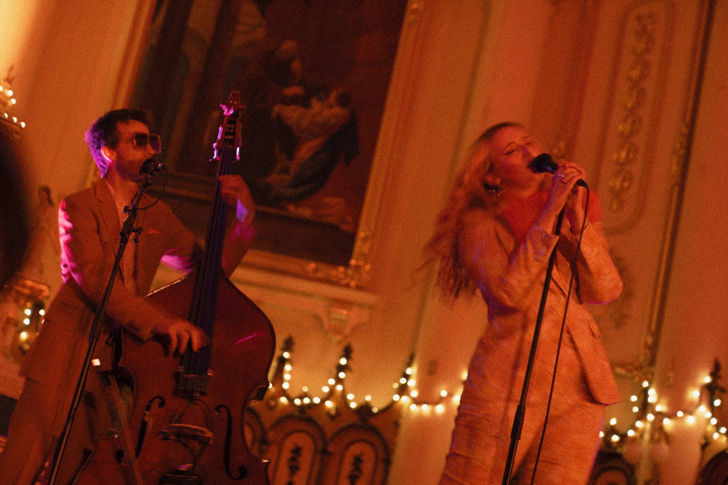 Deux musiciens, un homme jouant de la contrebasse et une femme chantant, se produisent dans une salle décorée de lumières chaudes et de peintures sur les murs.
