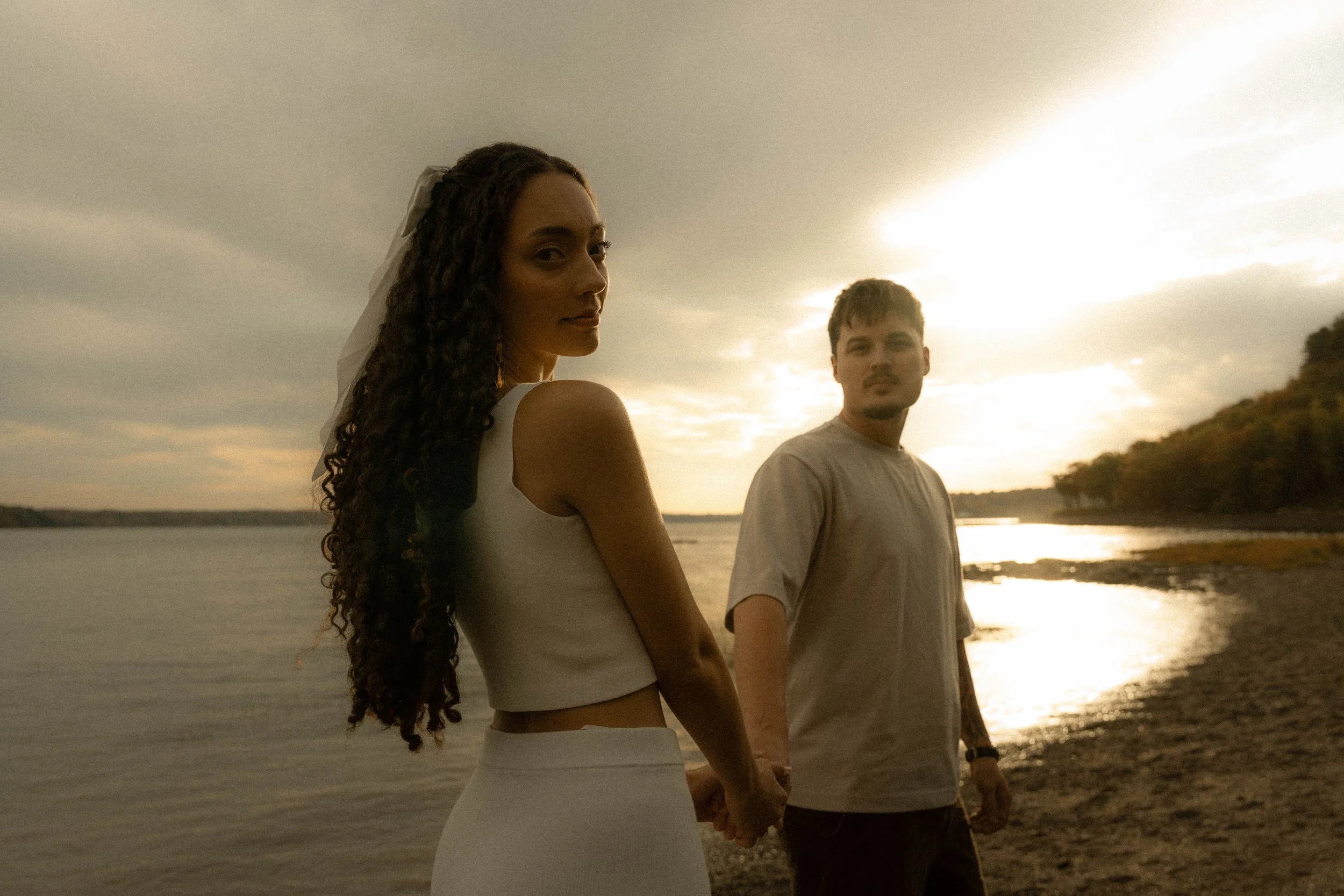 Un couple tenant une main se promène sur une plage au crépuscule, vue de dos. La femme a de longs cheveux bouclés et porte une robe blanche ou de couleur claire. L'homme a des cheveux courts et porte un t-shirt gris clair. Le soleil se couche derrièr