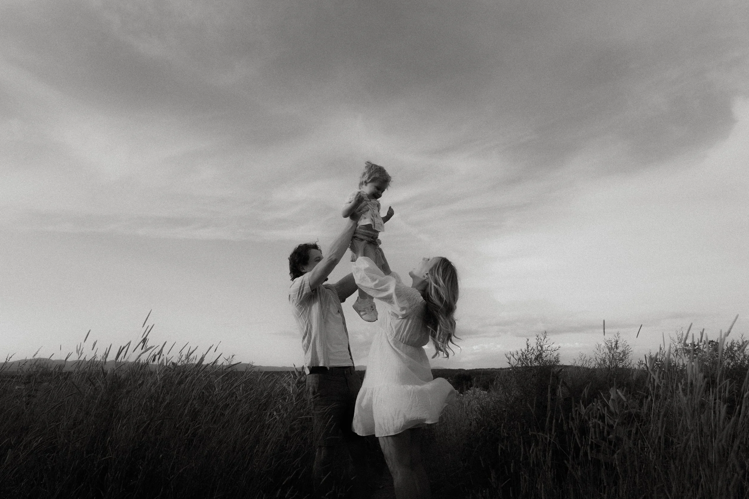 Une famille souriante dans un champ, deux adultes et une enfant, la fille est tenue en l'air par un adulte. Le ciel est nuageux et l'image est en noir et blanc.