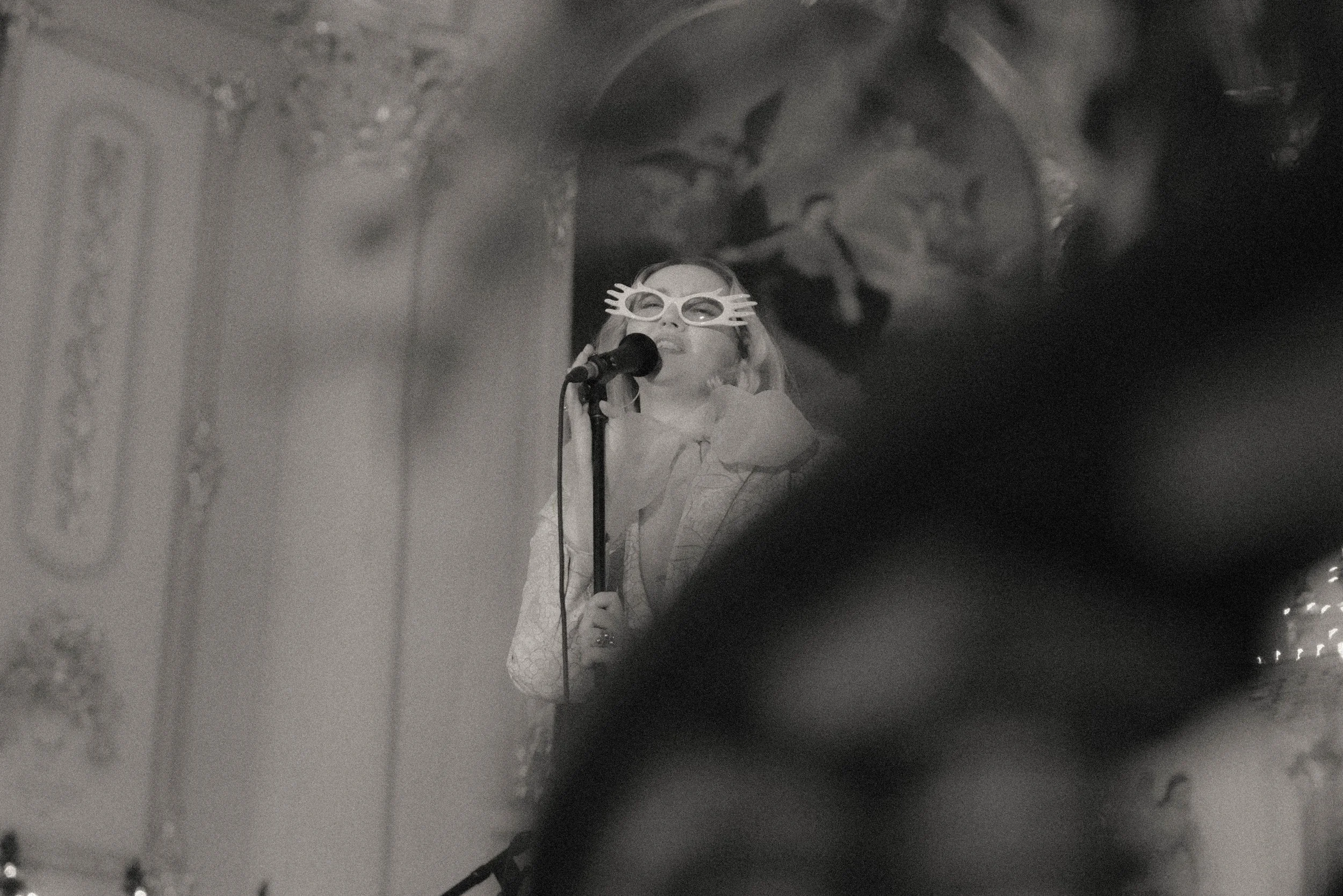 Une jeune fille portant des lunettes amusantes chante dans un microphone lors d'une performance sur scène, avec un décor élaboré en arrière-plan, en noir et blanc.