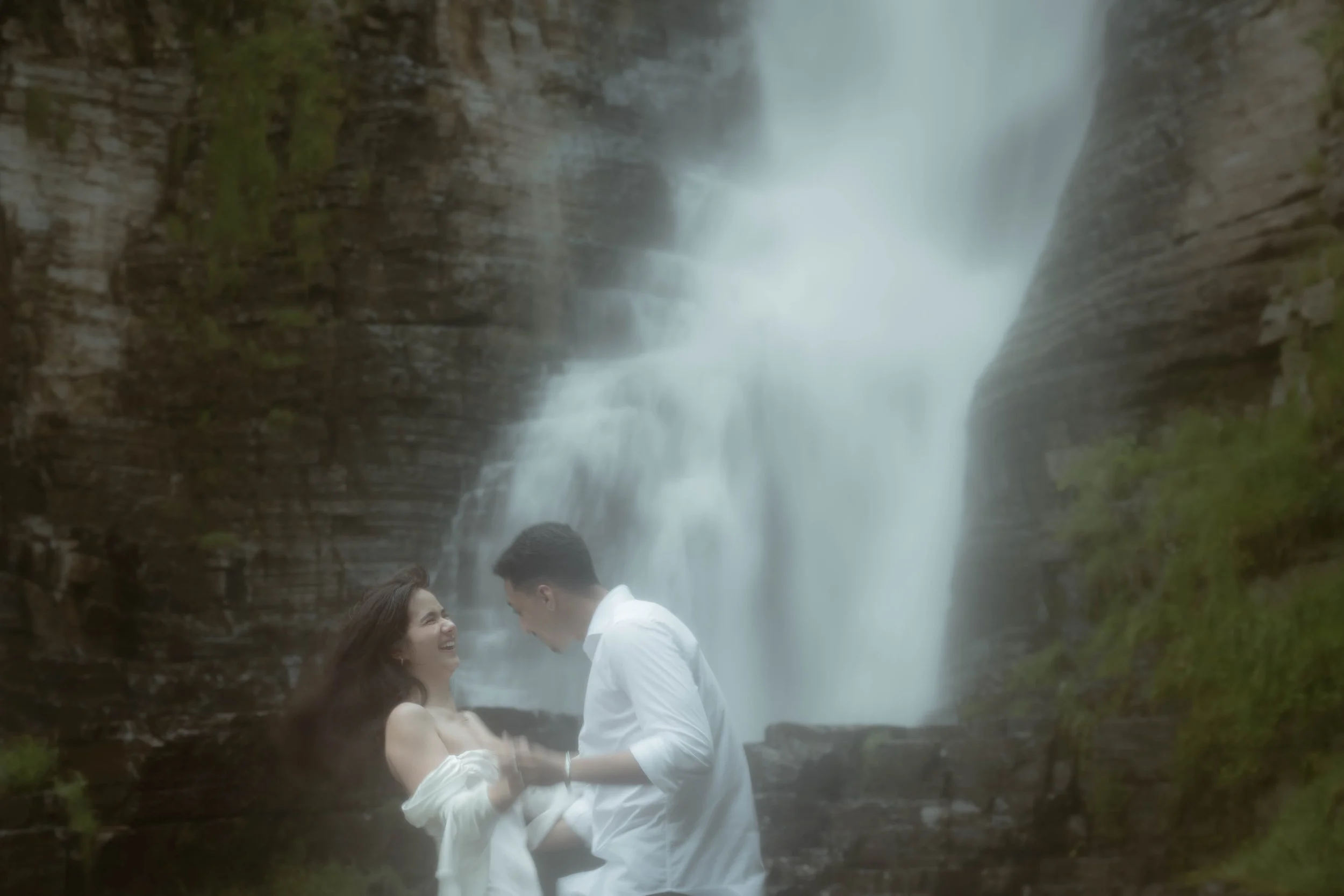 Un couple se tenant par la main sous une chute d'eau naturelle, souriant et regardant dans les yeux, dans un environnement forestier.