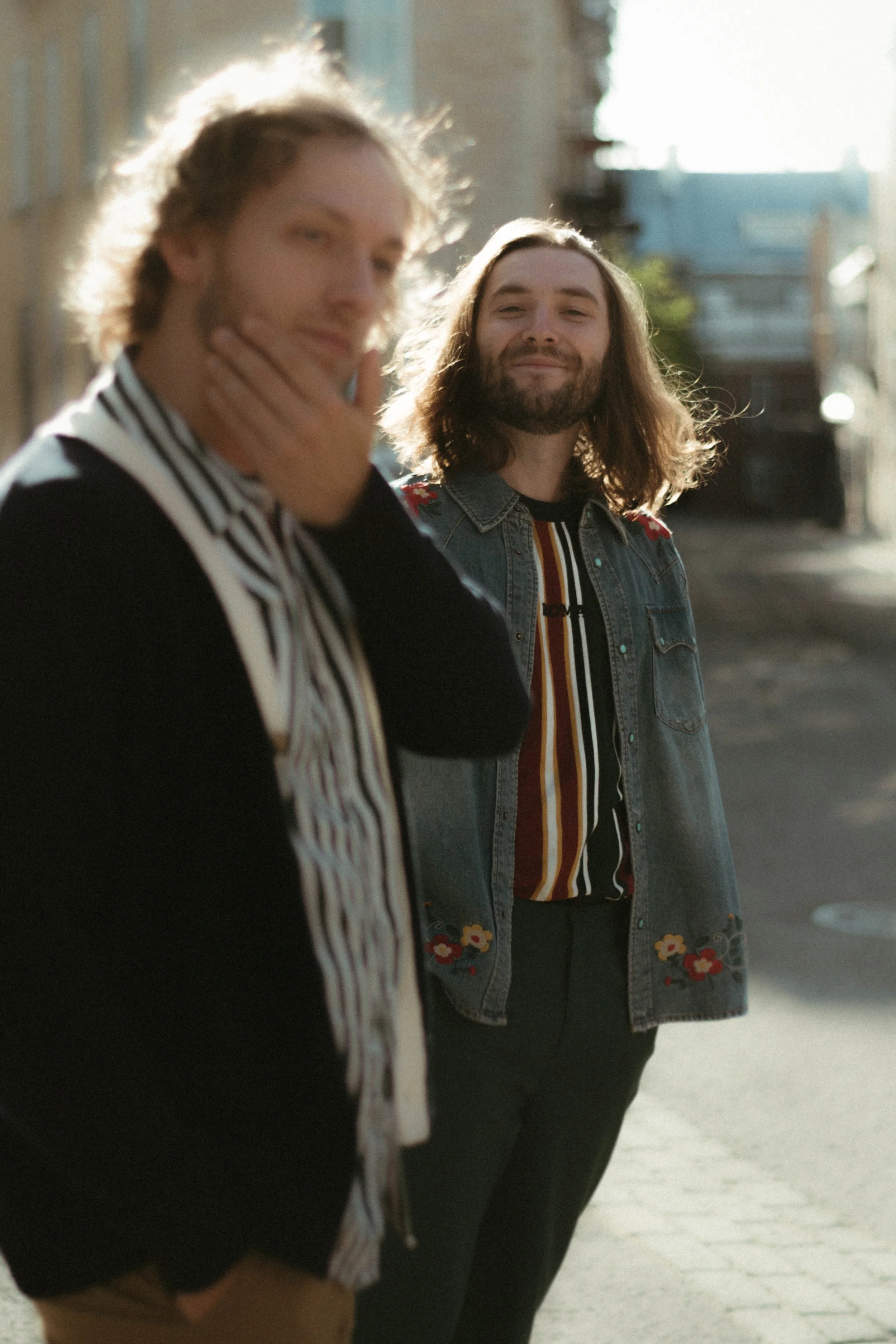 Deux personnes debout dans la rue, l'une en premier plan avec un chandail noir à rayures blanches, la seconde en arrière-plan avec une veste en jean brodée de fleurs, souriant.