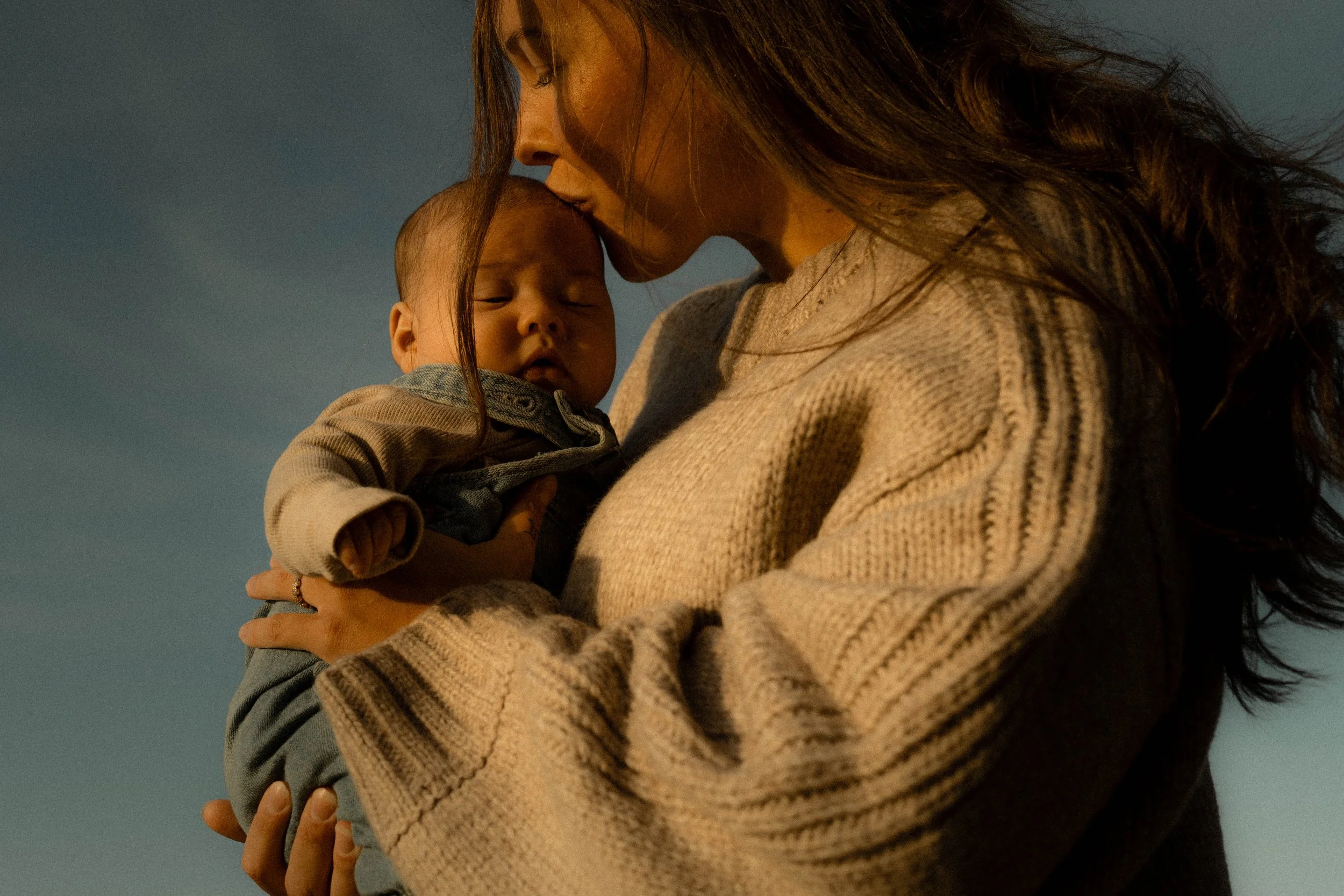 Une femme tient un bébé dans ses bras, elle lui donne un baiser sur le front. La femme porte un vêtement beige à rayures et a de longs cheveux bruns. Le bébé a des cheveux courts, porte une tenue gris clair et semble dormir.